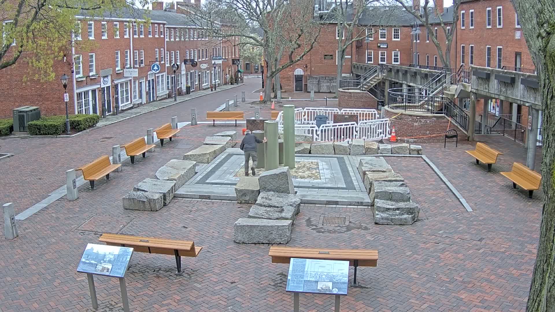 A person stands near a stone structure in a brick courtyard between two rows of brick buildings on an overcast day.
