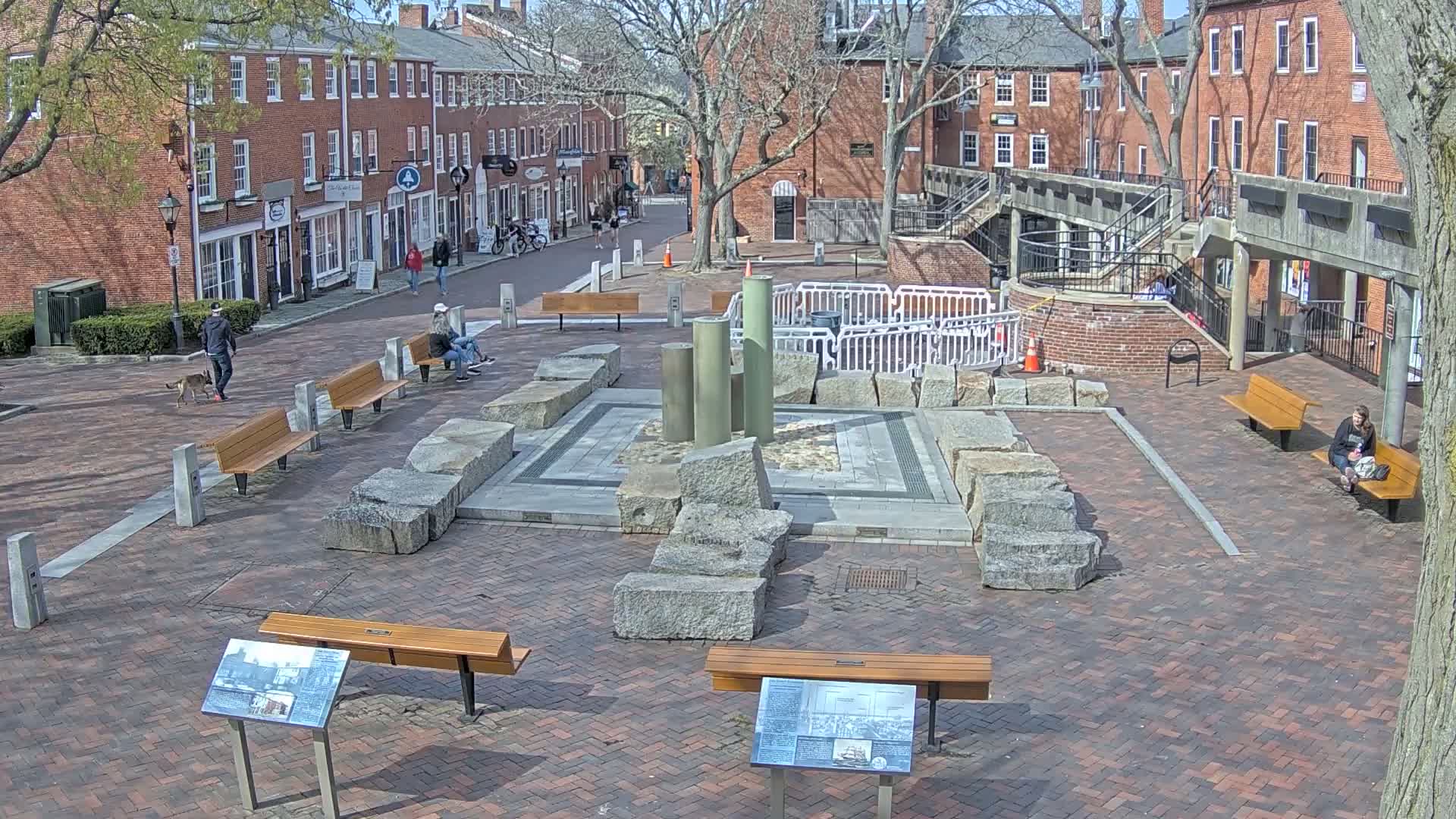 A brick plaza with several benches surrounds a stone structure featuring large granite blocks and tall columns, on a sunny day.