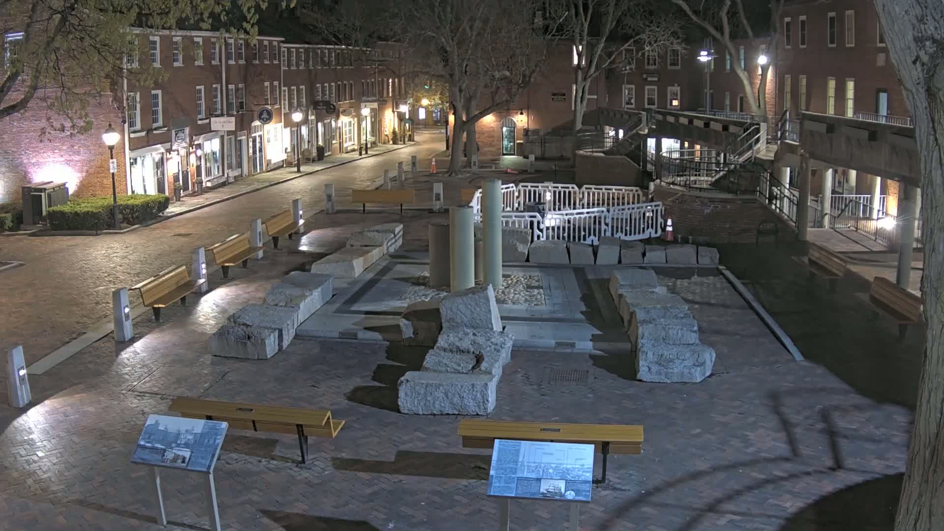 A nighttime view of a brick-paved plaza with large stone remnants, benches, and several buildings lining a street under a canopy of trees.