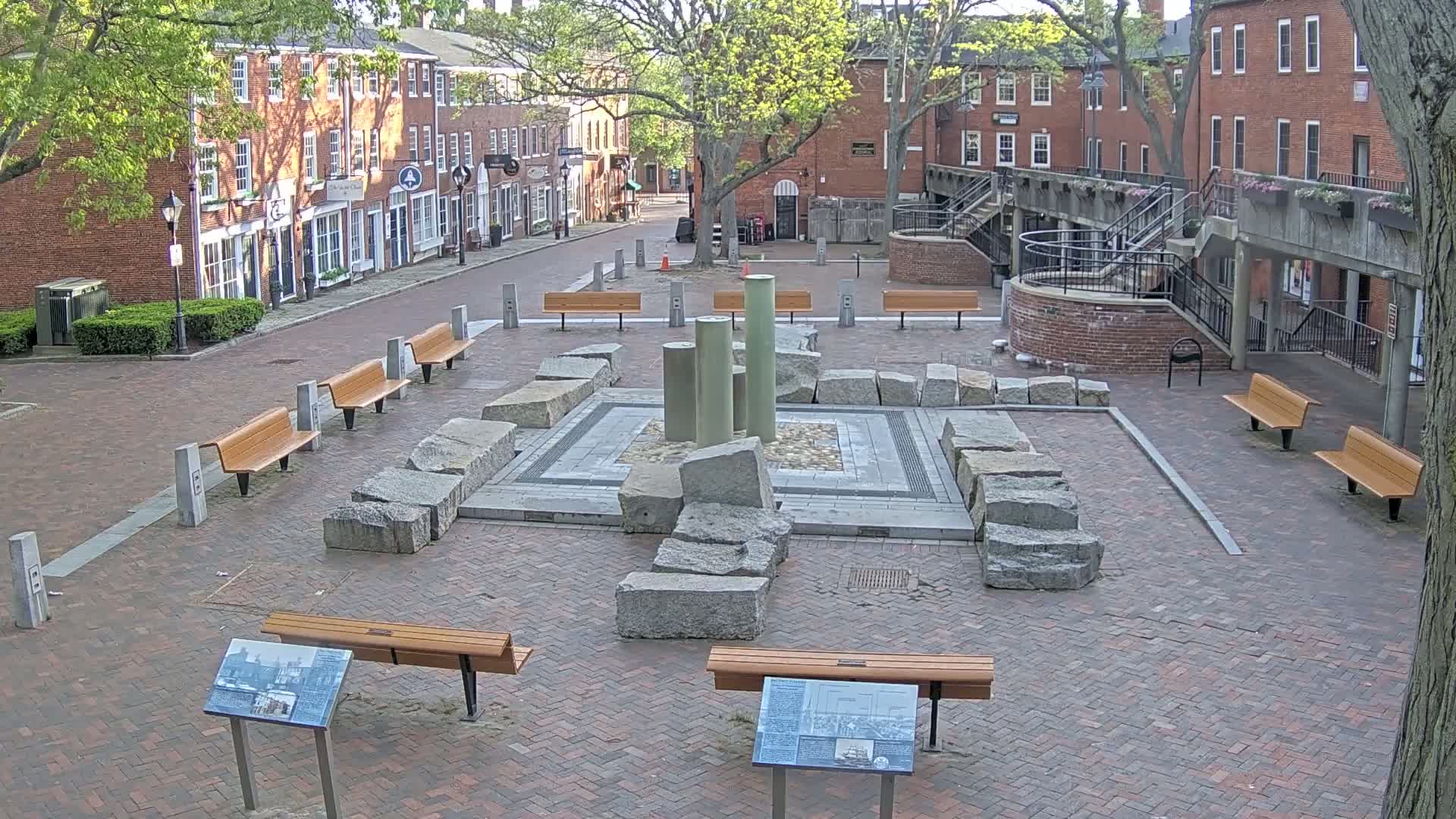 A brick plaza with several benches surrounds a stone fountain and several informational plaques, under a partly cloudy sky.