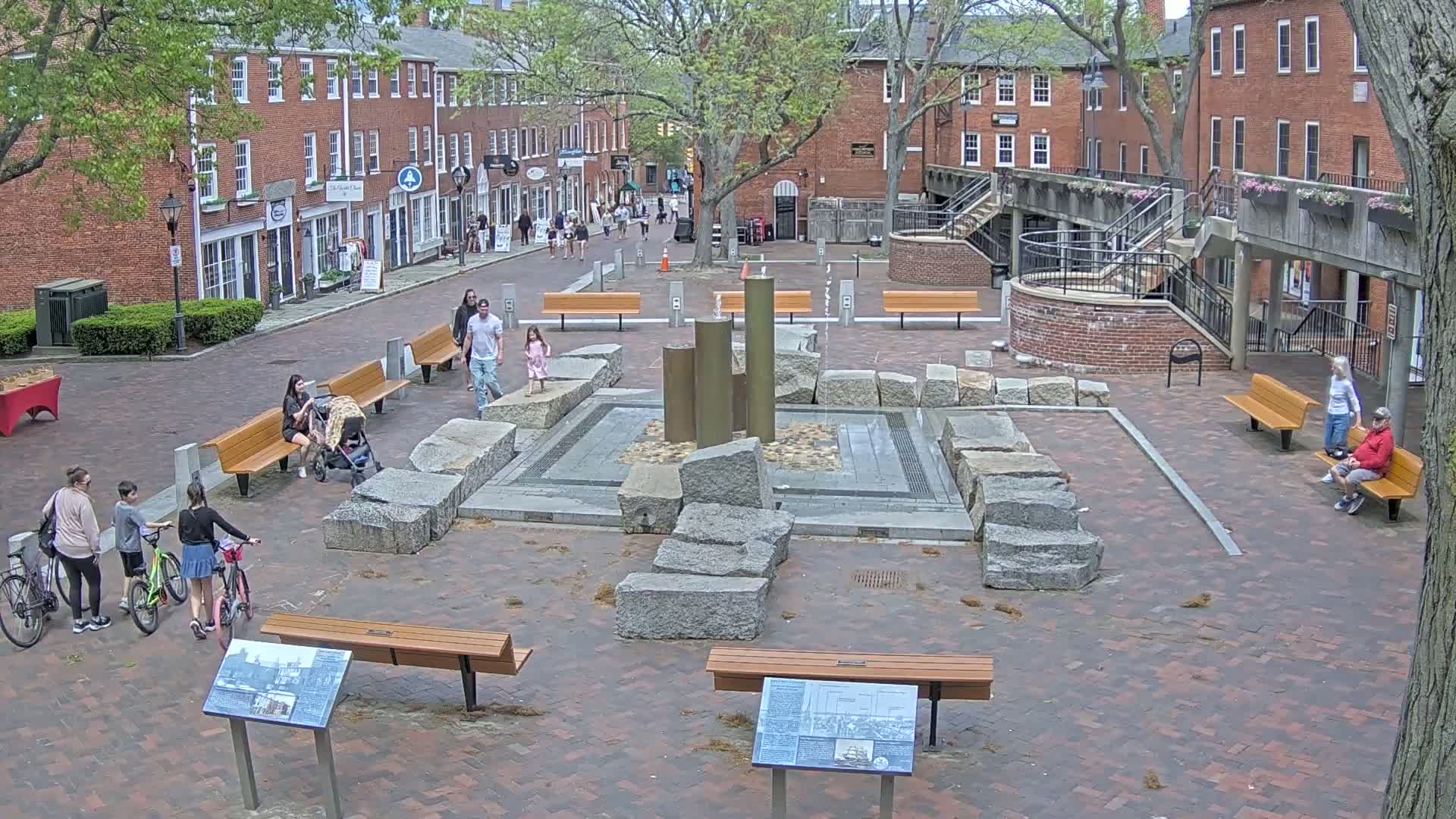 A brick plaza with a central fountain, benches, and several people strolling or sitting, under a partly sunny sky.