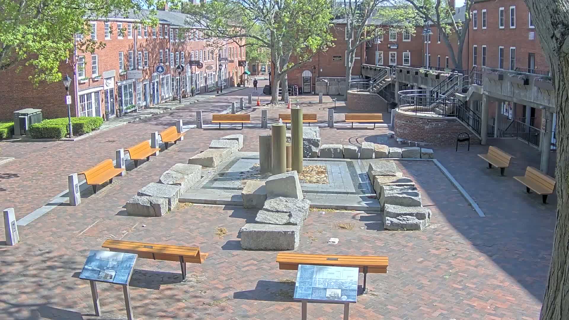 A brick plaza with a central fountain surrounded by large rocks, several benches, and information plaques, bordered by brick buildings under a sunny sky.
