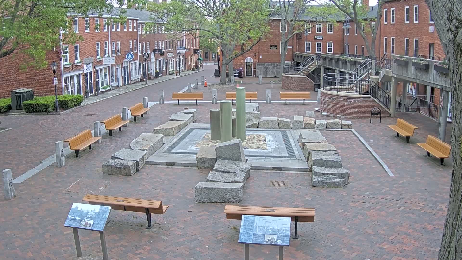 A brick plaza with benches, a central stone structure, and several brick buildings in the background on a partly cloudy day.