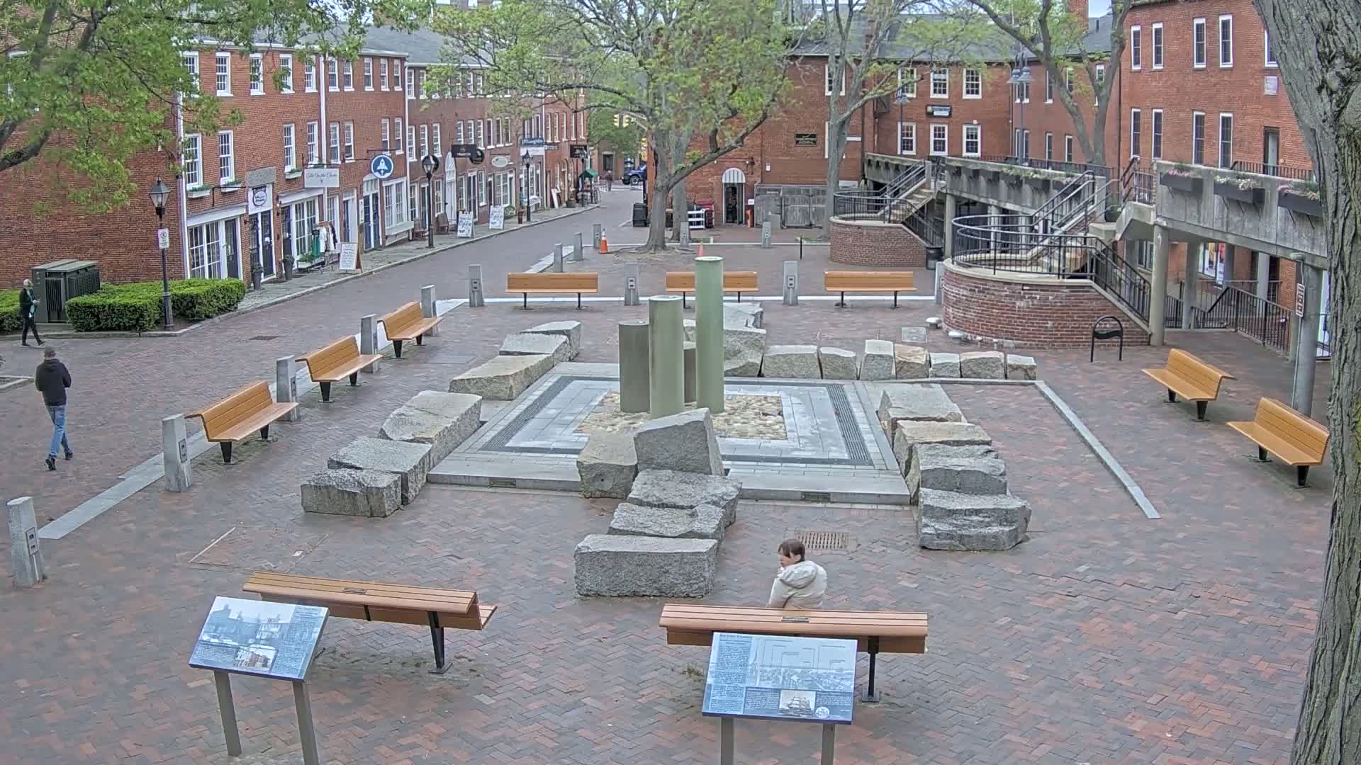 A brick plaza with several benches and a stone sculpture is surrounded by brick buildings under a partly cloudy sky.