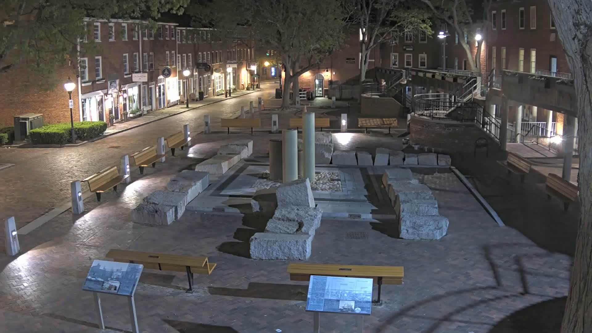 A nighttime view of a brick-paved plaza with several benches, a central stone structure resembling ancient ruins, and brick buildings illuminated by streetlights in the background.
