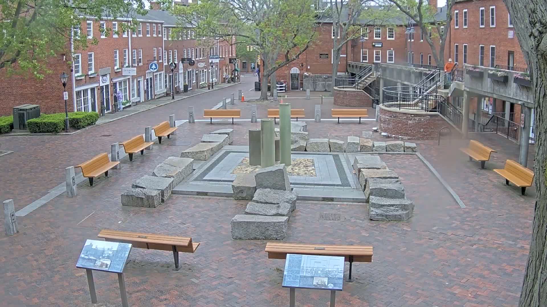 A brick plaza with several benches surrounds a central fountain-like structure made of large granite stones, all situated between brick buildings under an overcast sky.