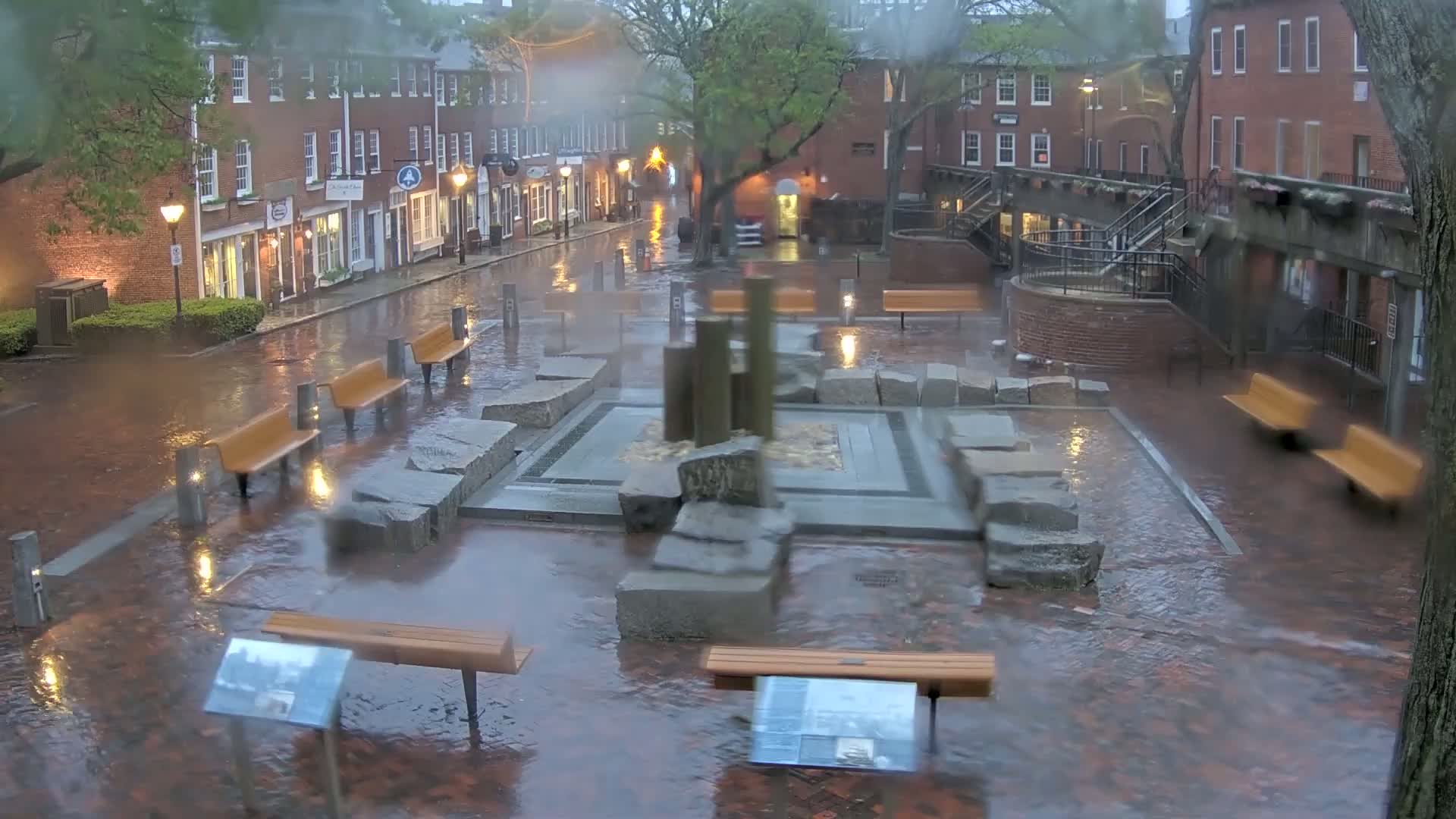 A rain-soaked brick plaza features a stone fountain and several benches under the gloomy light of street lamps, situated between buildings in a city.