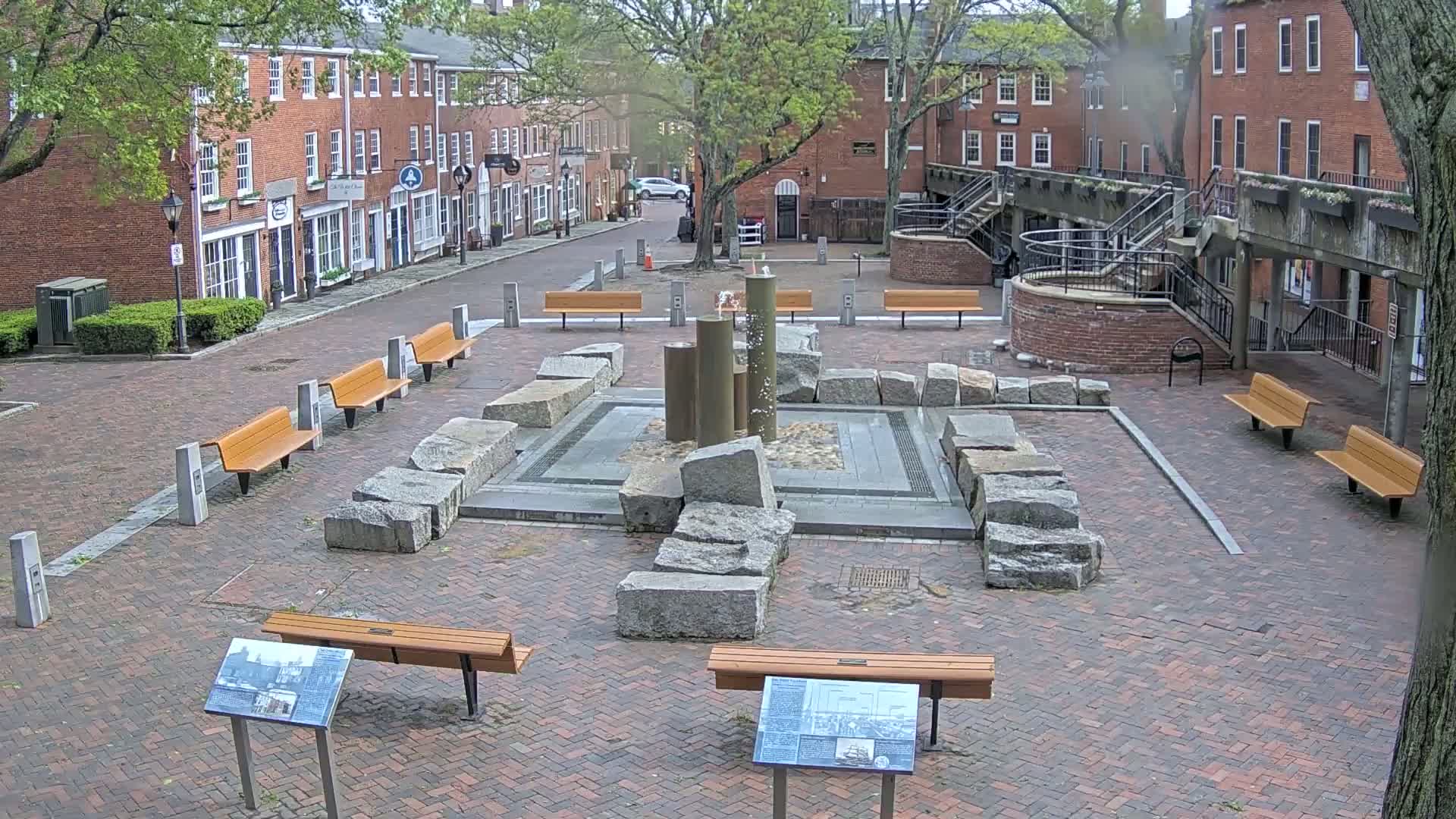 A brick plaza with a central fountain surrounded by large granite stones and several wooden benches, under an overcast sky.