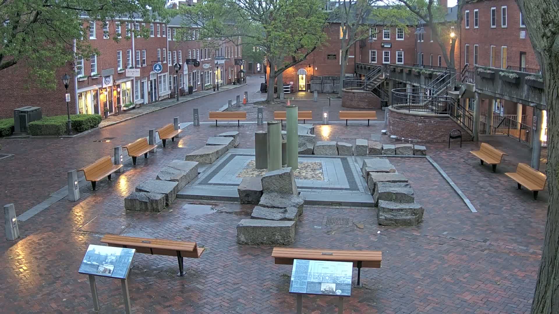 A wet brick plaza with several benches surrounds a stone fountain, situated between brick buildings under a cloudy sky.