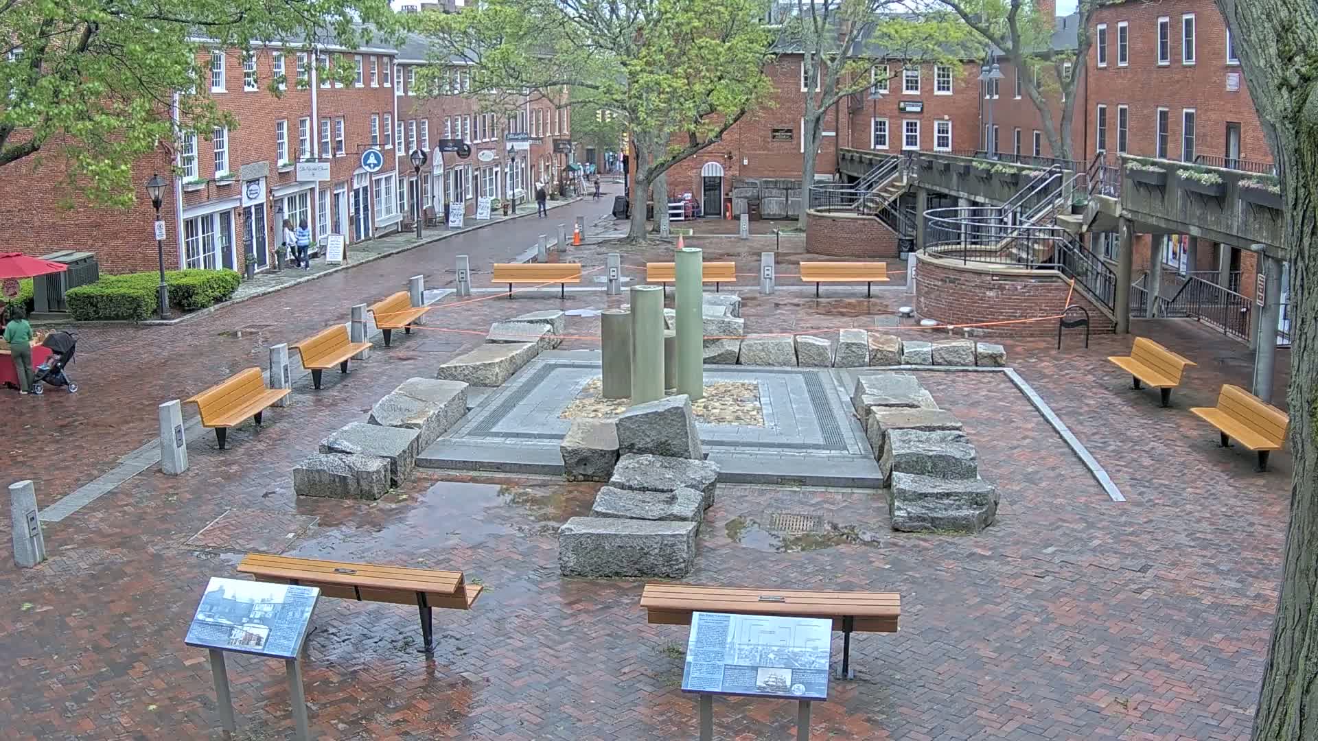 A brick plaza with several benches surrounds a stone and metal fountain, wet from recent rain, in front of brick buildings.