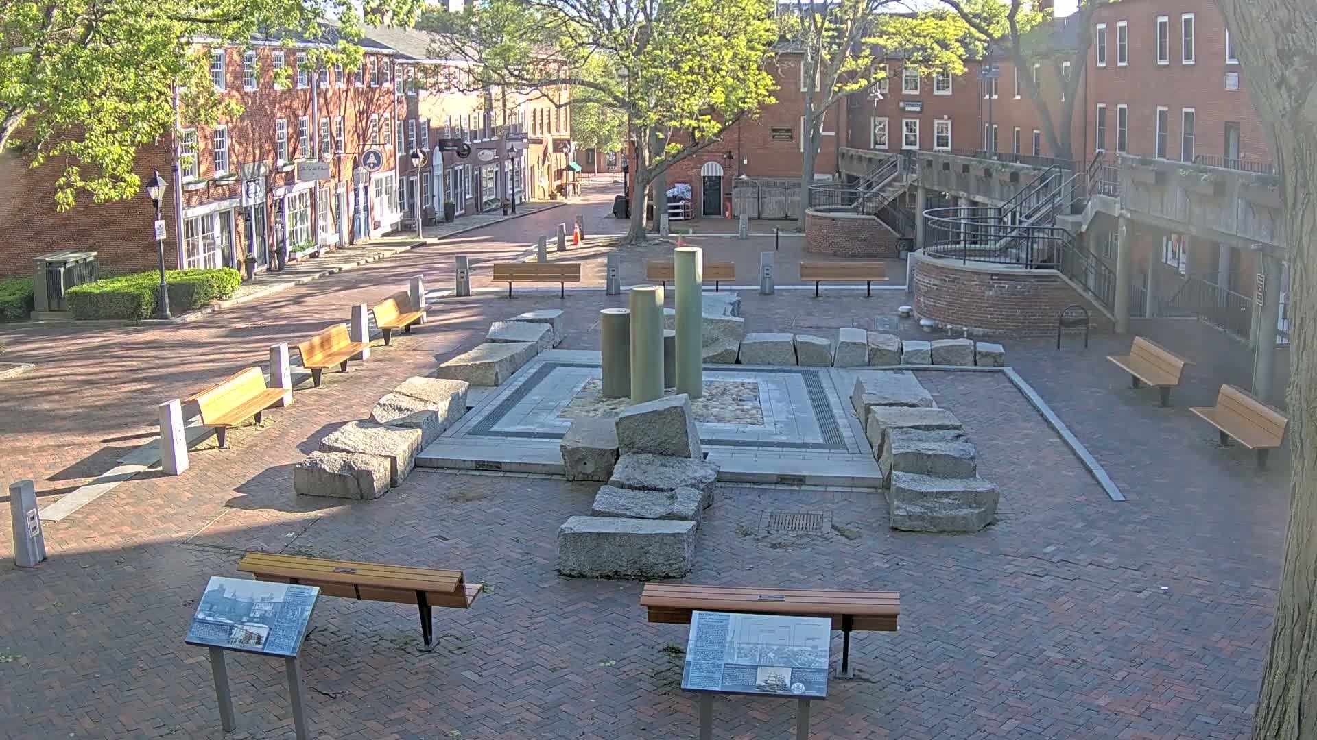 A brick plaza with several benches surrounds a stone monument featuring several upright columns, under a partly sunny sky.