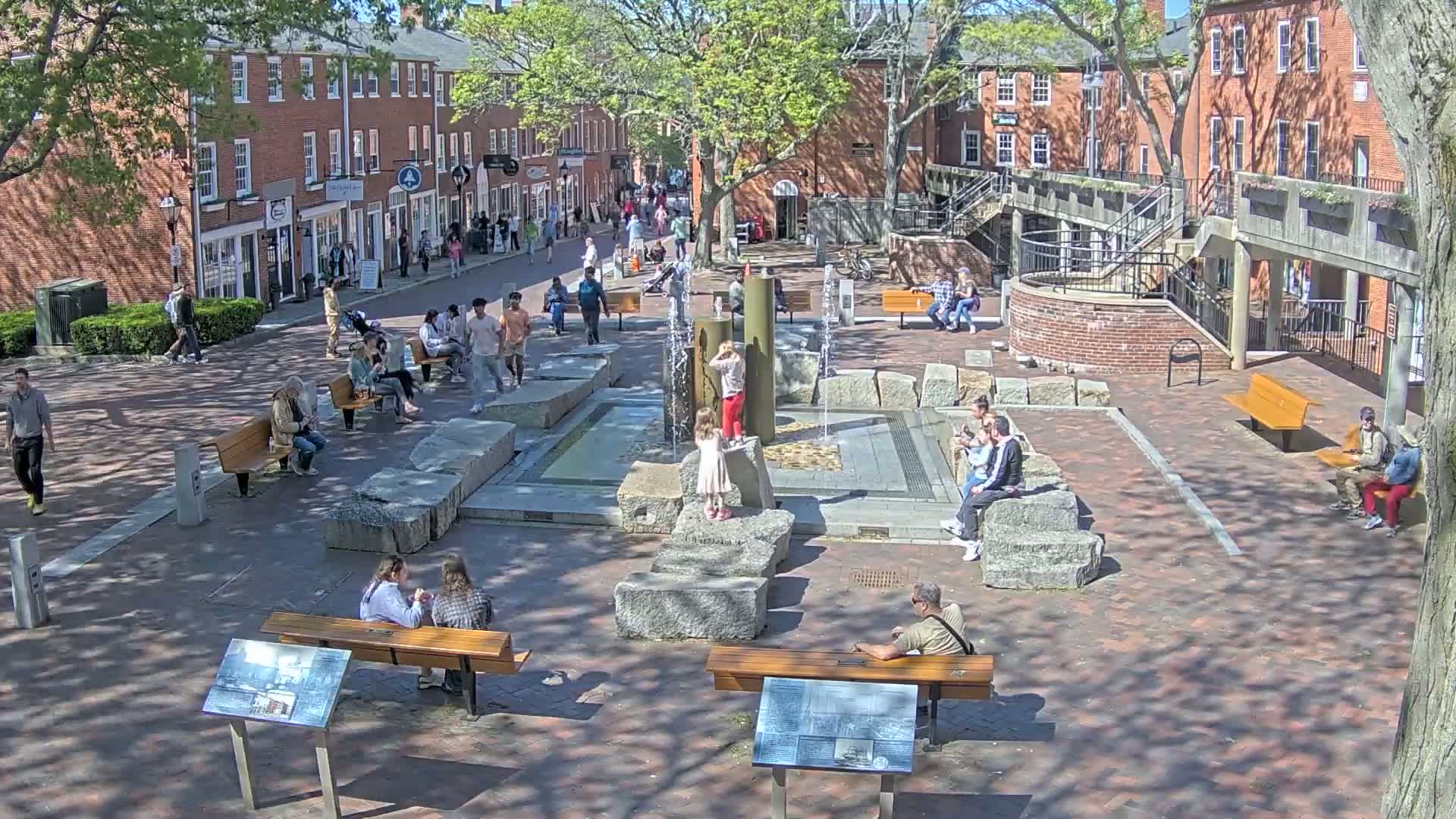 A sunny town square features a central fountain surrounded by people sitting on benches and large rocks, with brick buildings lining the square.