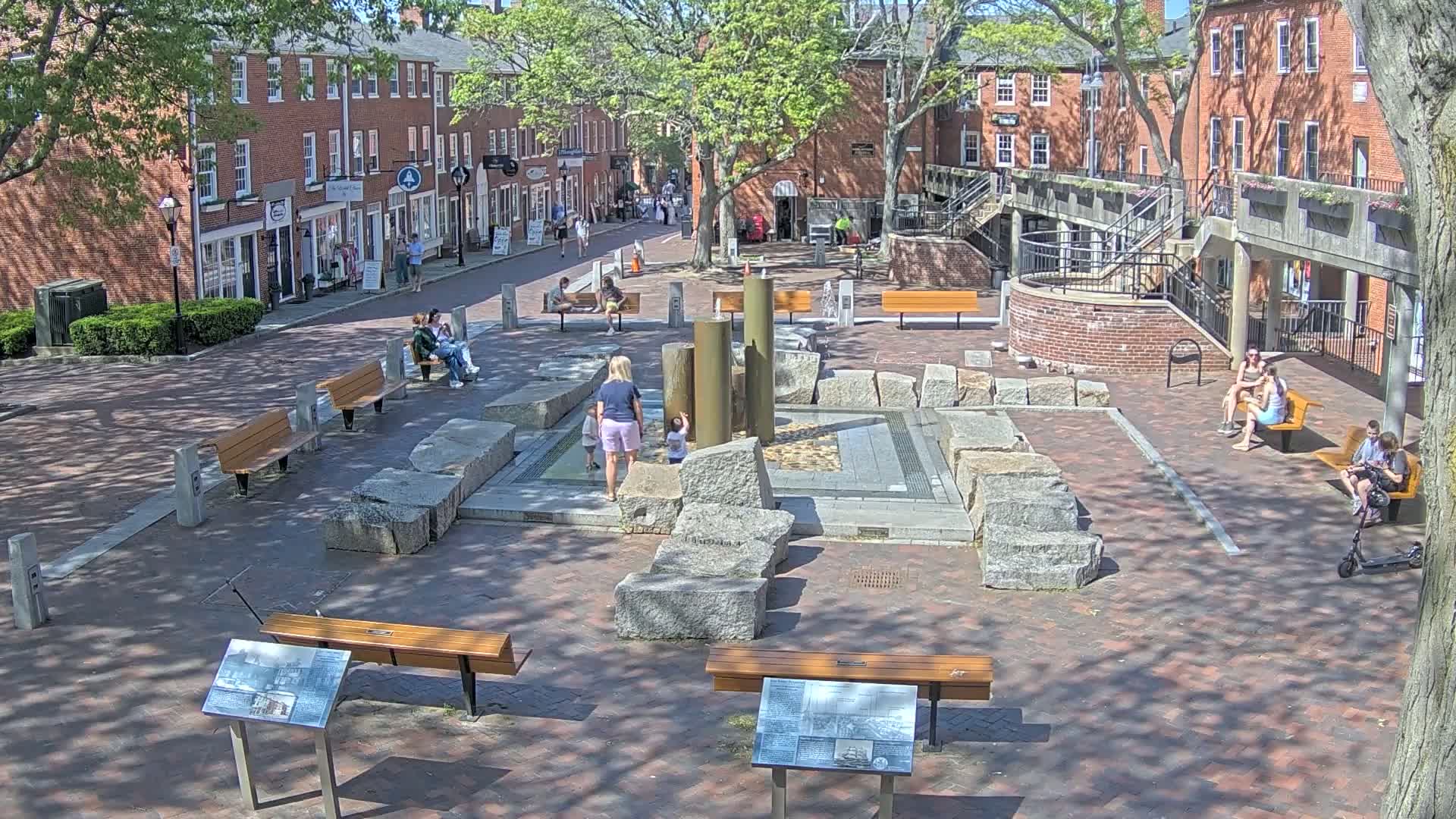 A sunny day in a brick-paved town square, featuring a central water fountain surrounded by large stones, benches, and people sitting and playing.