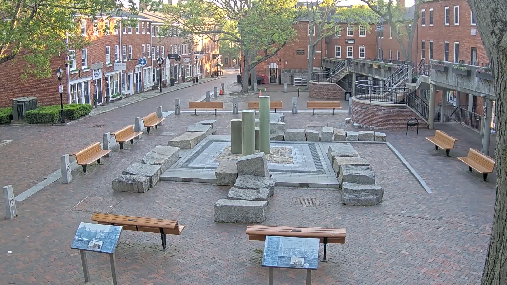 A brick plaza features a stone and metal fountain surrounded by benches and several informational plaques, with brick buildings and trees visible in the background under a sunny sky.