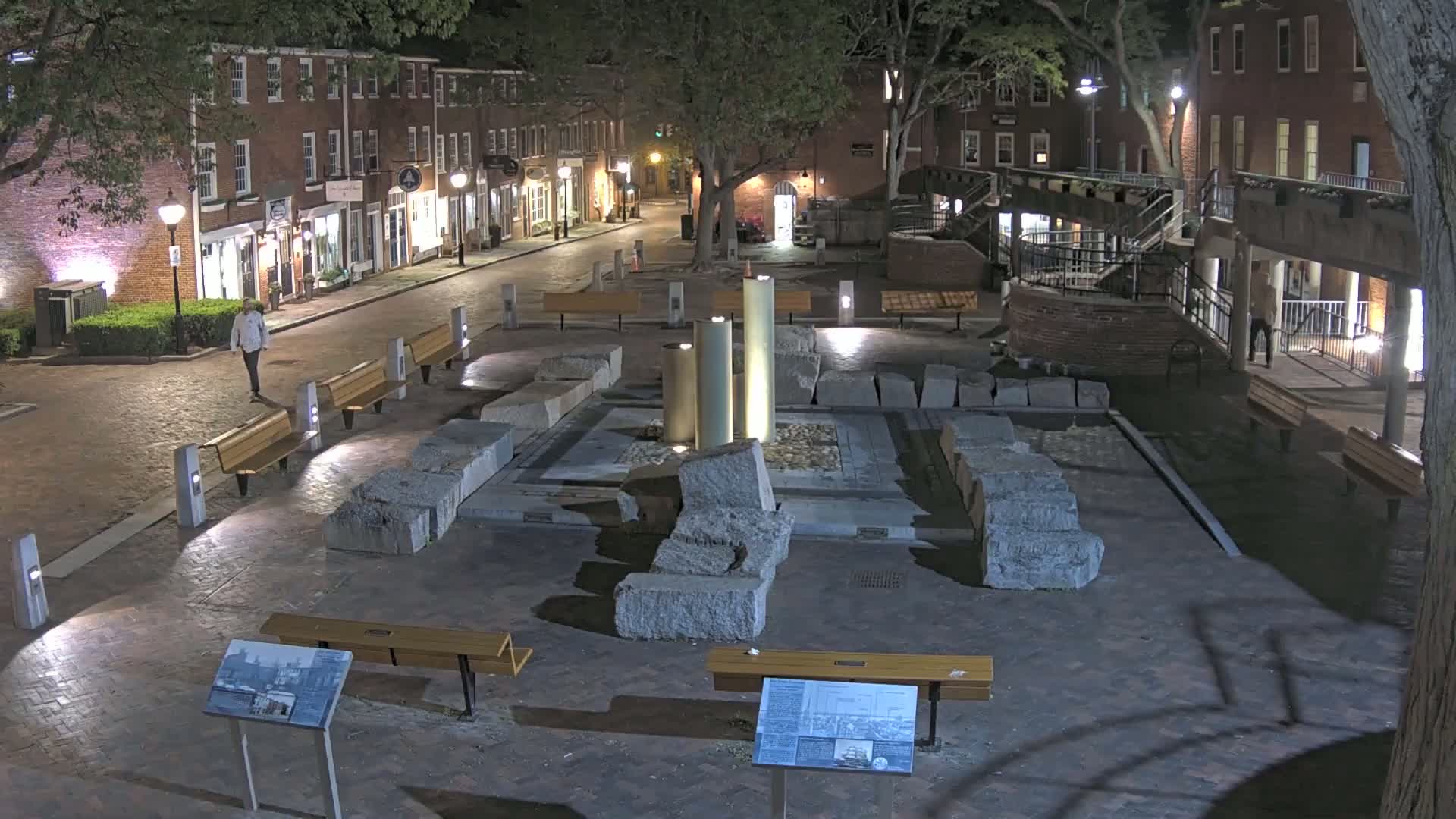 At night, a brick-lined plaza features a central fountain surrounded by large stone blocks, with benches and several buildings lining the street.