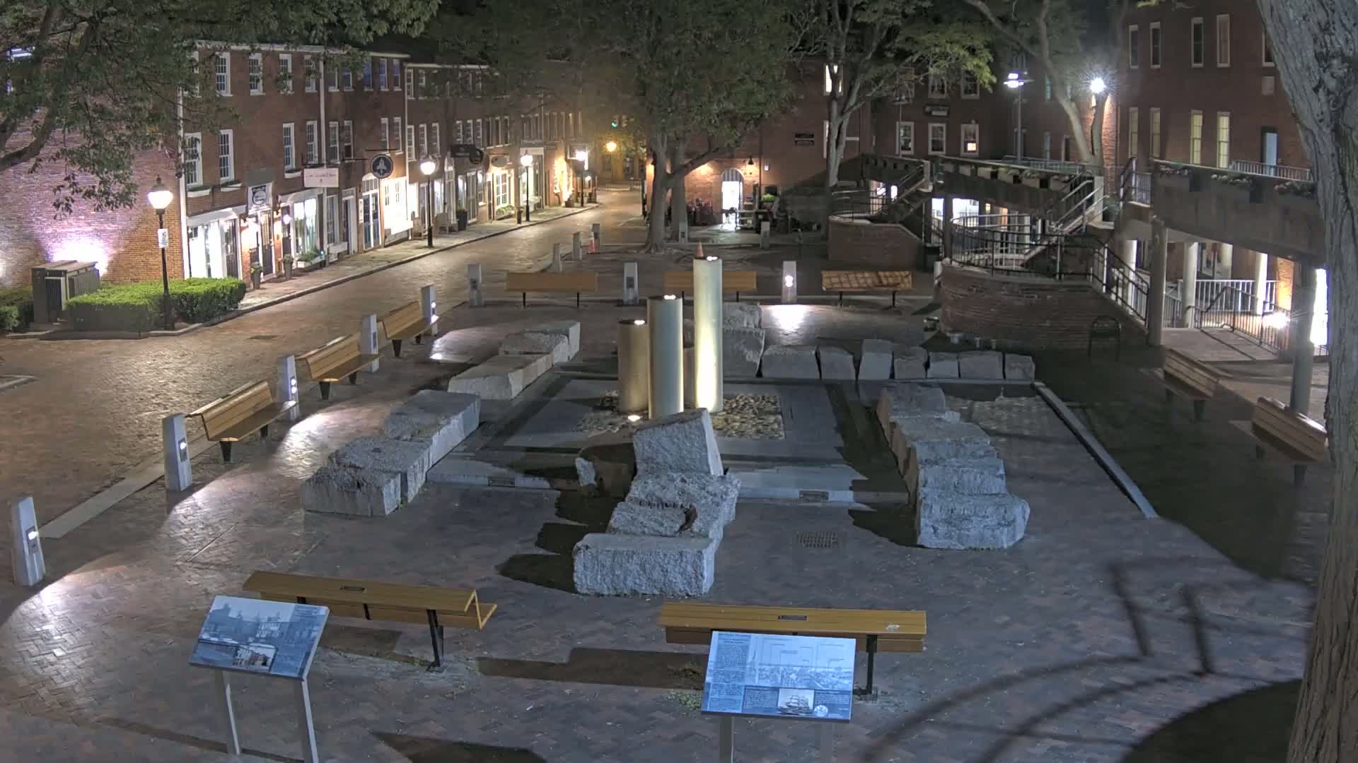 At night, a brick-lined street with shops and benches leads to a plaza featuring a stone structure and several illuminated cylindrical pillars.