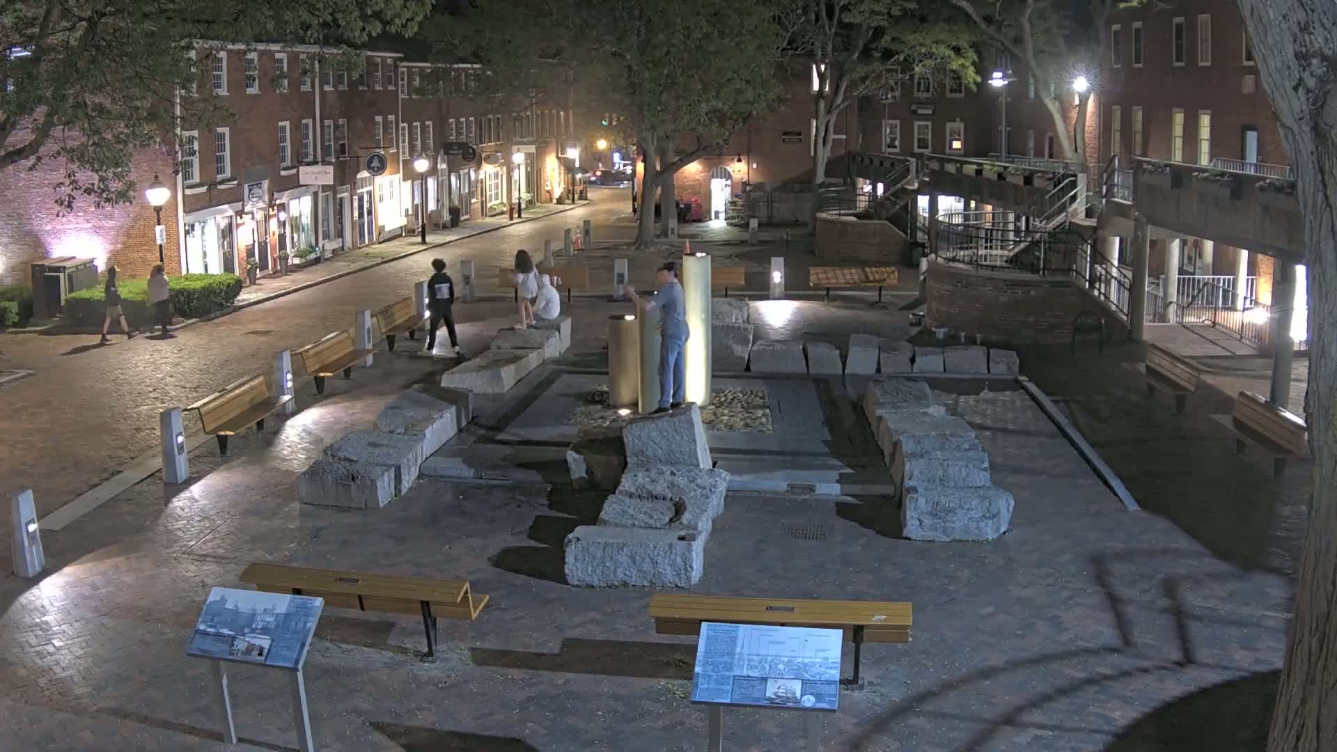 At night, a brick-paved plaza with several people, benches, and a stone structure is illuminated by streetlights.