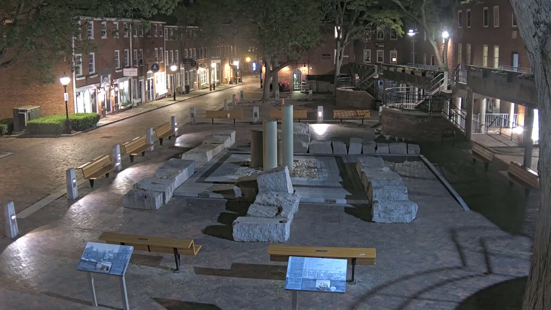 At night, a brick-lined plaza features a stone ruin in the center, surrounded by benches and illuminated by streetlights.