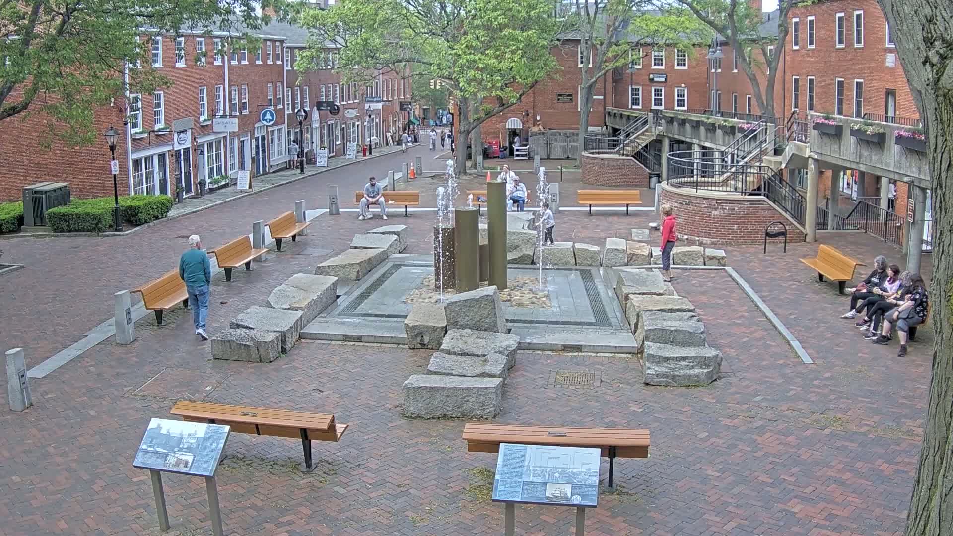 A brick plaza with a fountain in the center, surrounded by benches, several people, and brick buildings under a partly cloudy sky.