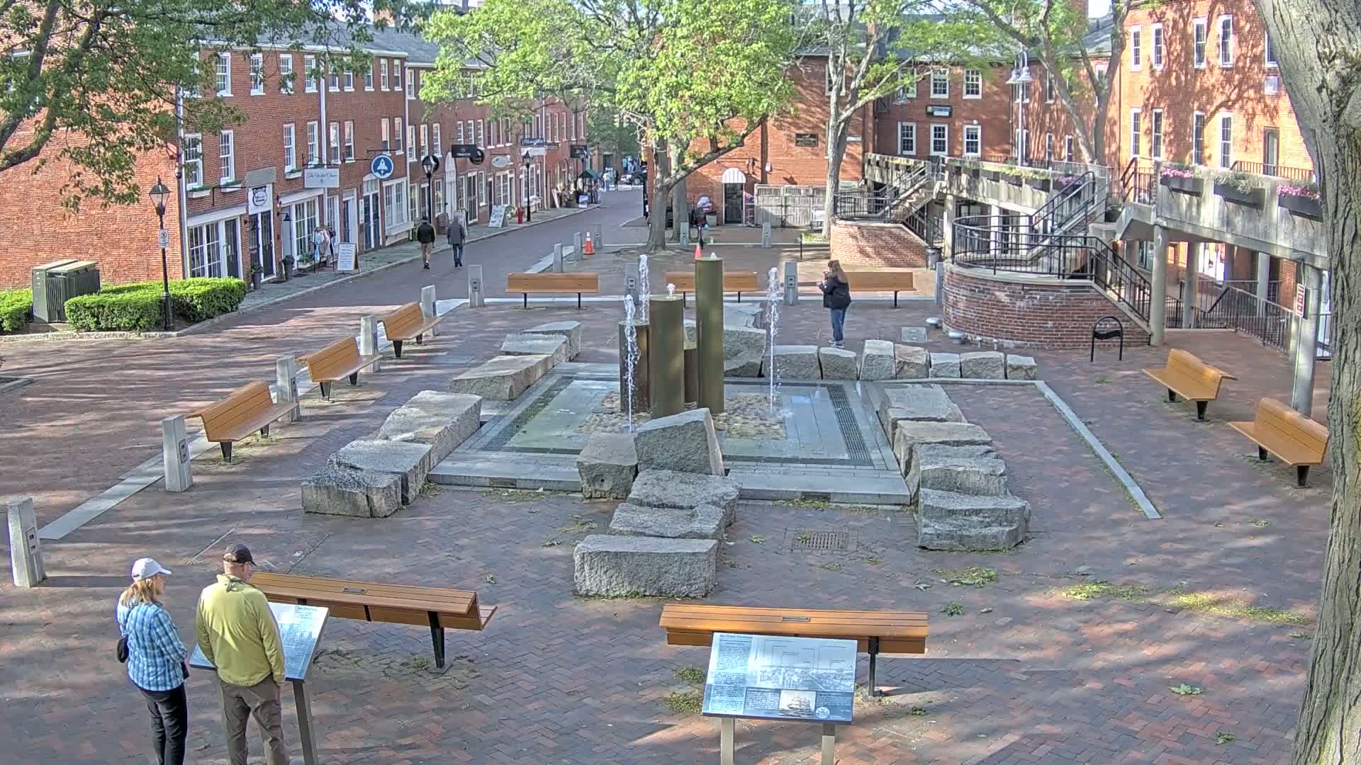 A brick plaza with a fountain surrounded by large stones and wooden benches, situated between brick buildings on a sunny day.