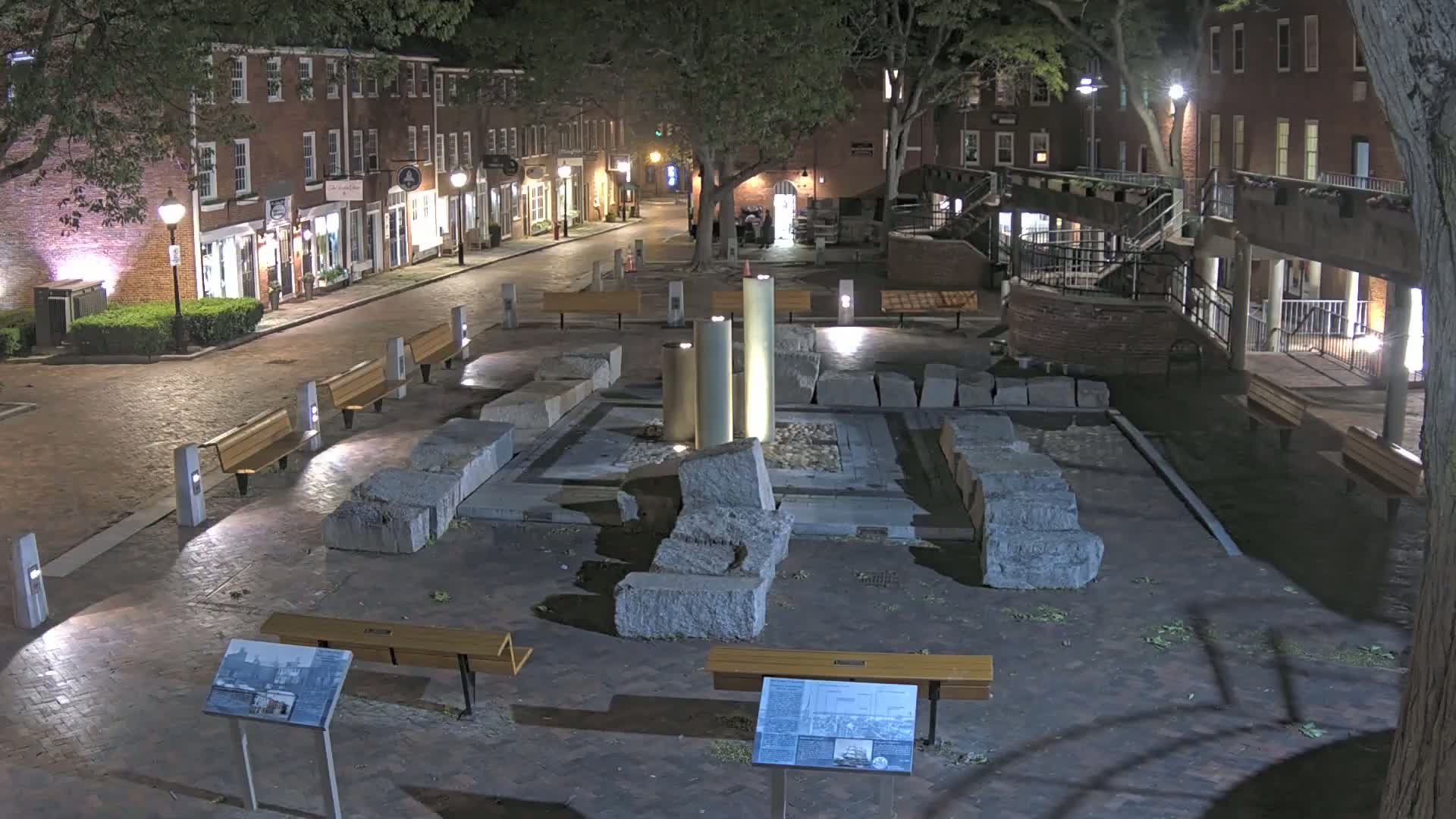 A nighttime view of a brick paved plaza with a central stone fountain structure, surrounded by benches and lit by streetlights, situated between brick buildings under a canopy of trees.