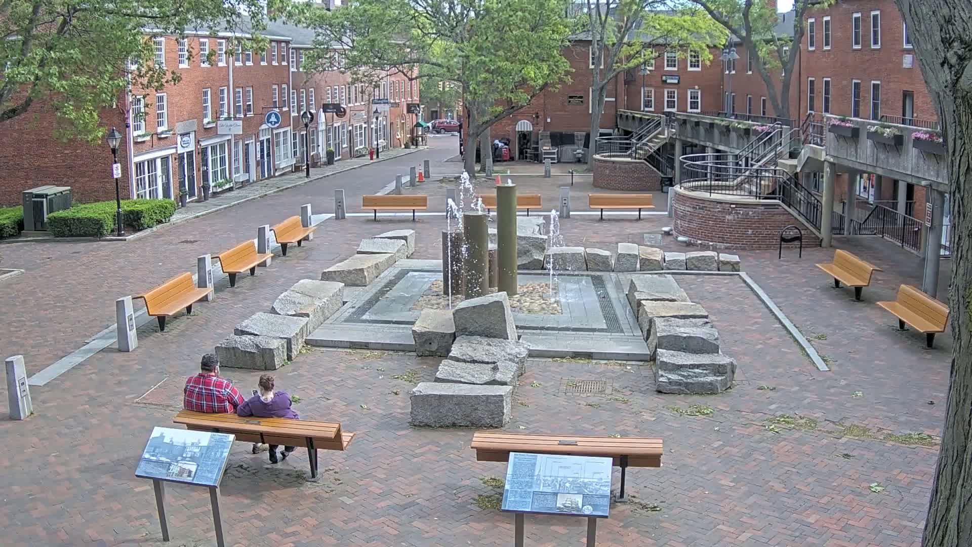 A brick plaza with a fountain surrounded by large granite rocks and several wooden benches, situated between brick buildings under a partly cloudy sky.