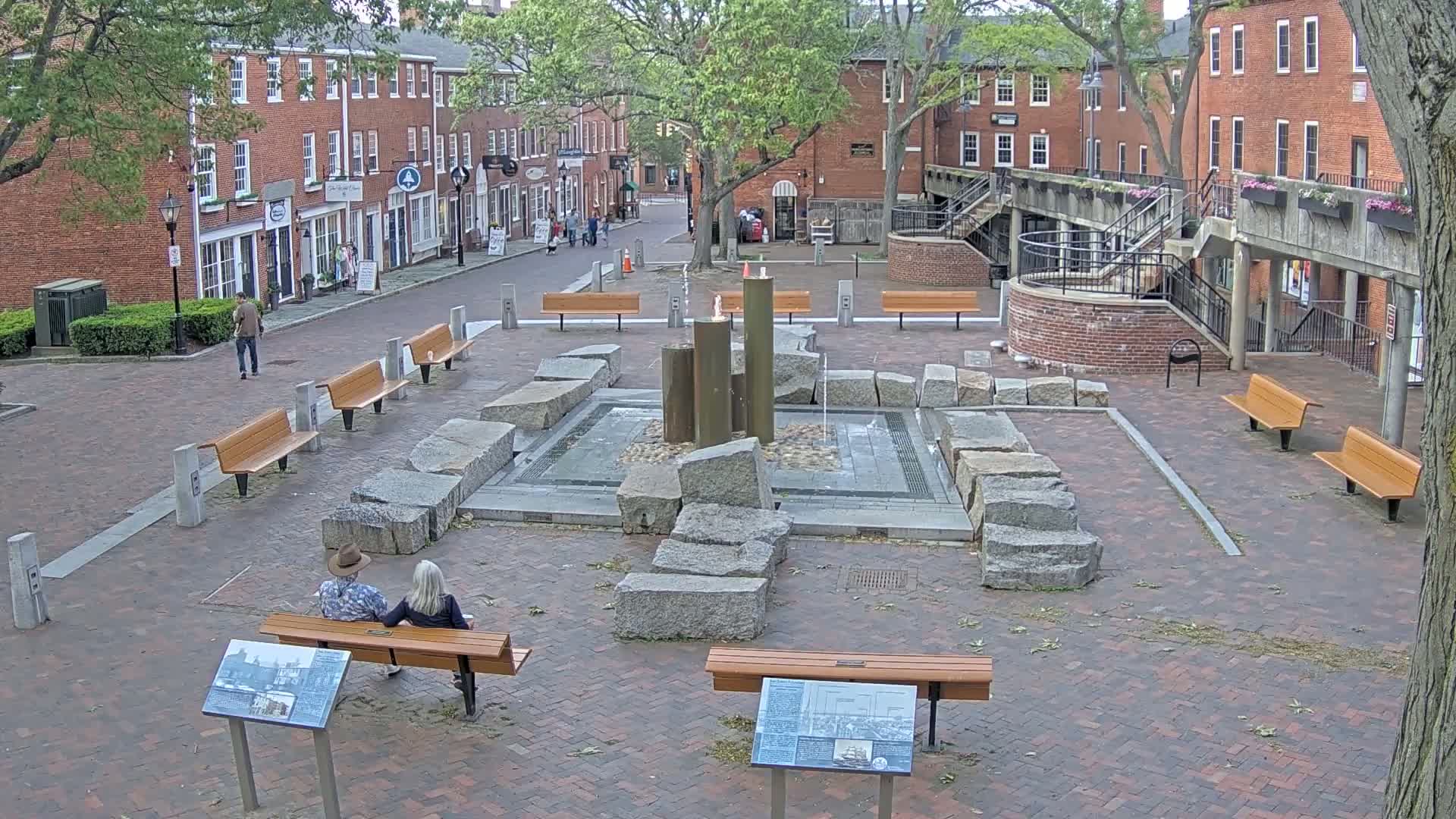 A brick town square with a stone fountain, benches, and brick buildings surrounds it under a partly cloudy sky.