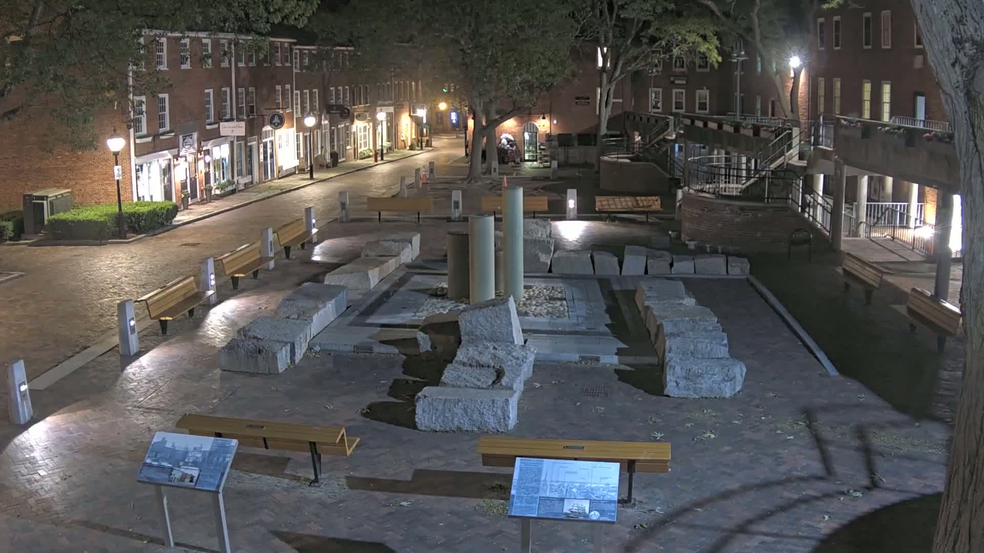 At night, a stone plaza with remnants of columns and several benches sits in the center of a cobblestone street lined with brick buildings under the shade of trees.