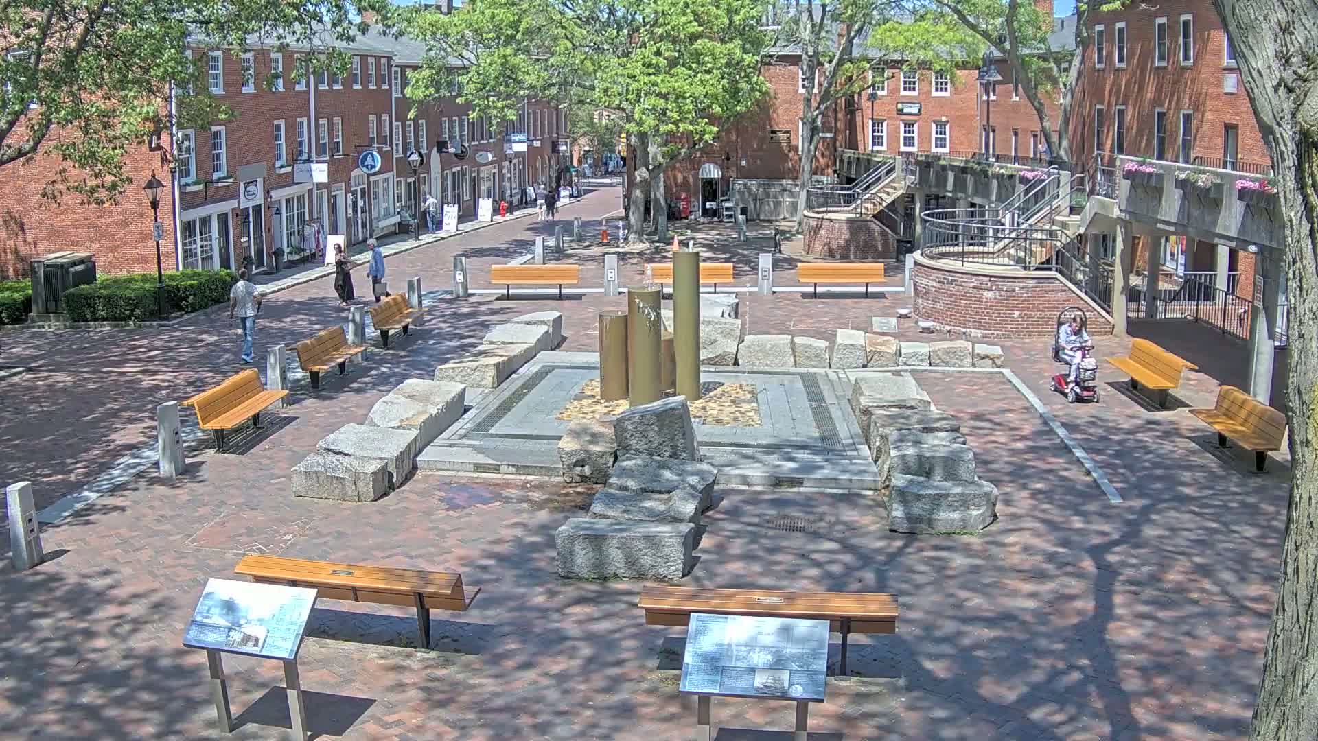 A brick plaza with a central fountain, several benches, and surrounding brick buildings on a sunny day.