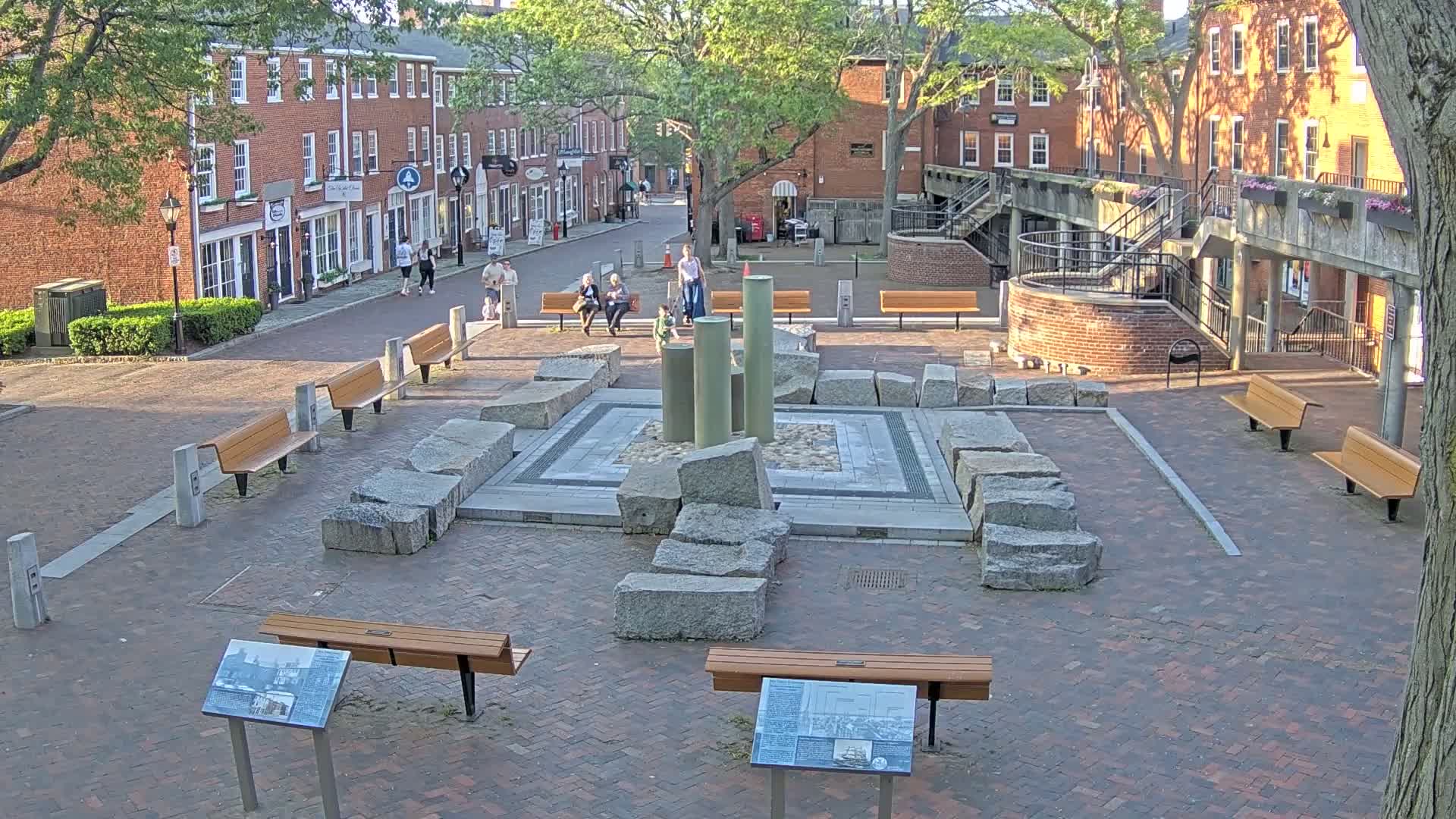 A brick plaza with several benches and a stone fountain is situated between two-story brick buildings under a partly sunny sky.