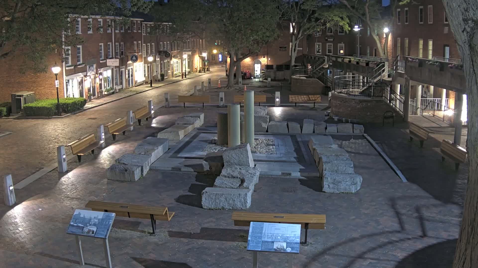 A nighttime view of a brick-paved town square featuring a central display of large stone blocks and columns, surrounded by benches and lit by street lamps.