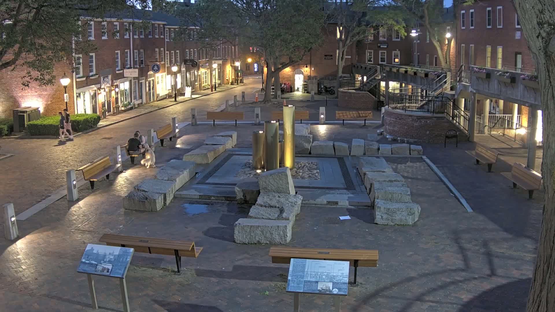 A nighttime scene shows a town square with a modern fountain surrounded by large stones and benches, situated between brick buildings under a mostly clear sky.
