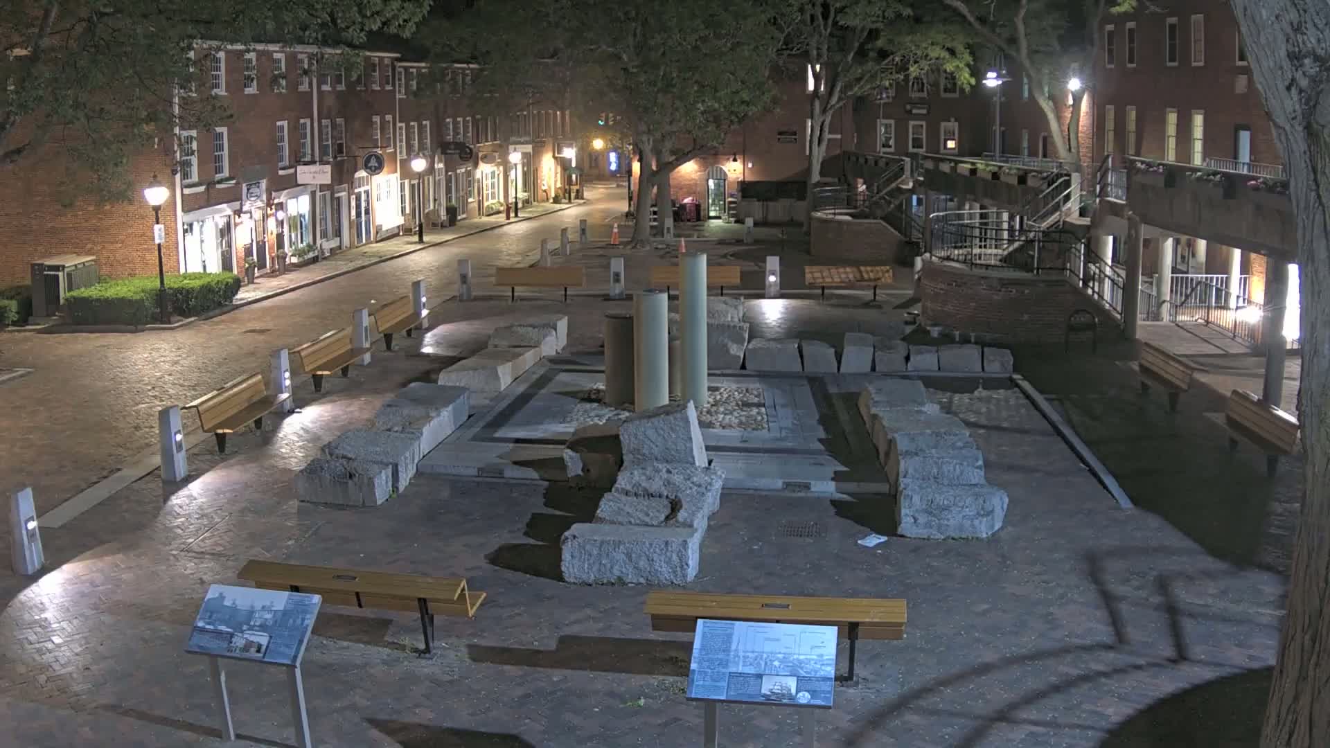 A nighttime view of a brick-paved plaza with stone ruins in the center, surrounded by benches, streetlights, and brick buildings.