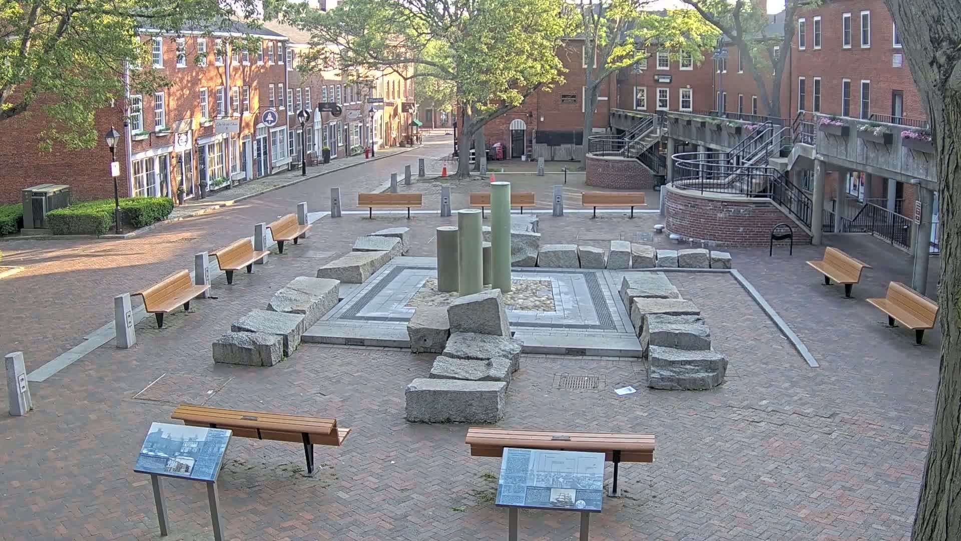 A brick plaza with several benches surrounds a central fountain composed of large granite stones and tall, pale green columns; the scene is outdoors under a partly sunny sky.