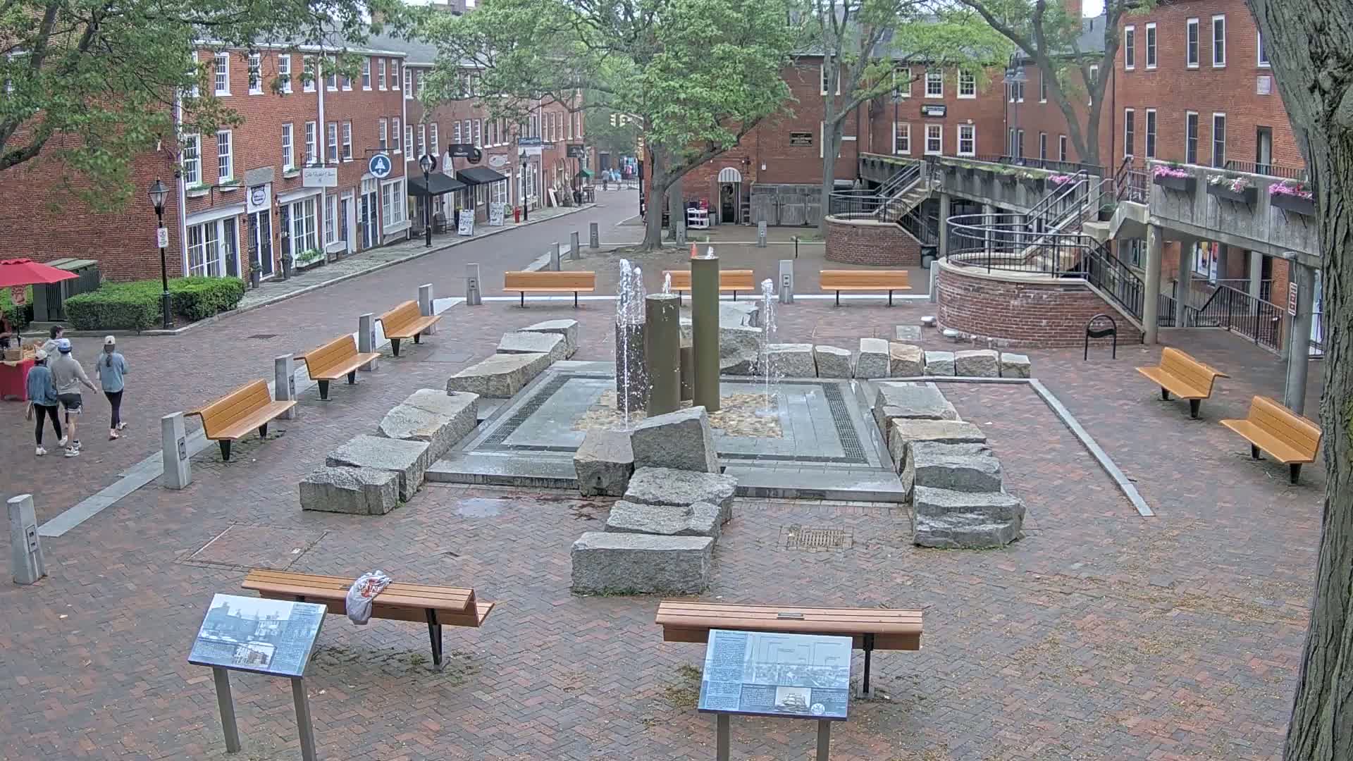 A brick plaza with a central fountain surrounded by benches and several brick buildings under an overcast sky.