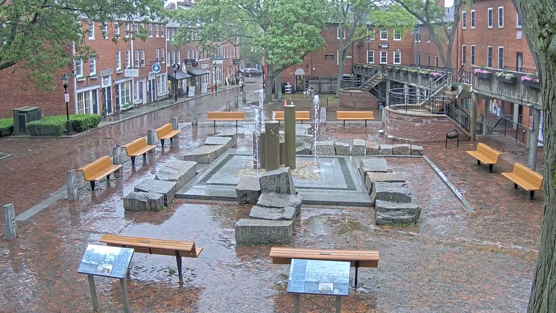 A wet brick plaza features a fountain surrounded by large stones and several benches, with rain-slicked streets and buildings visible in the background.