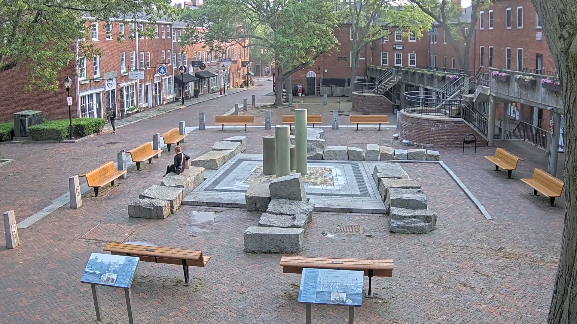 A brick courtyard with several benches surrounds a stone fountain-like structure, under a partly cloudy sky.