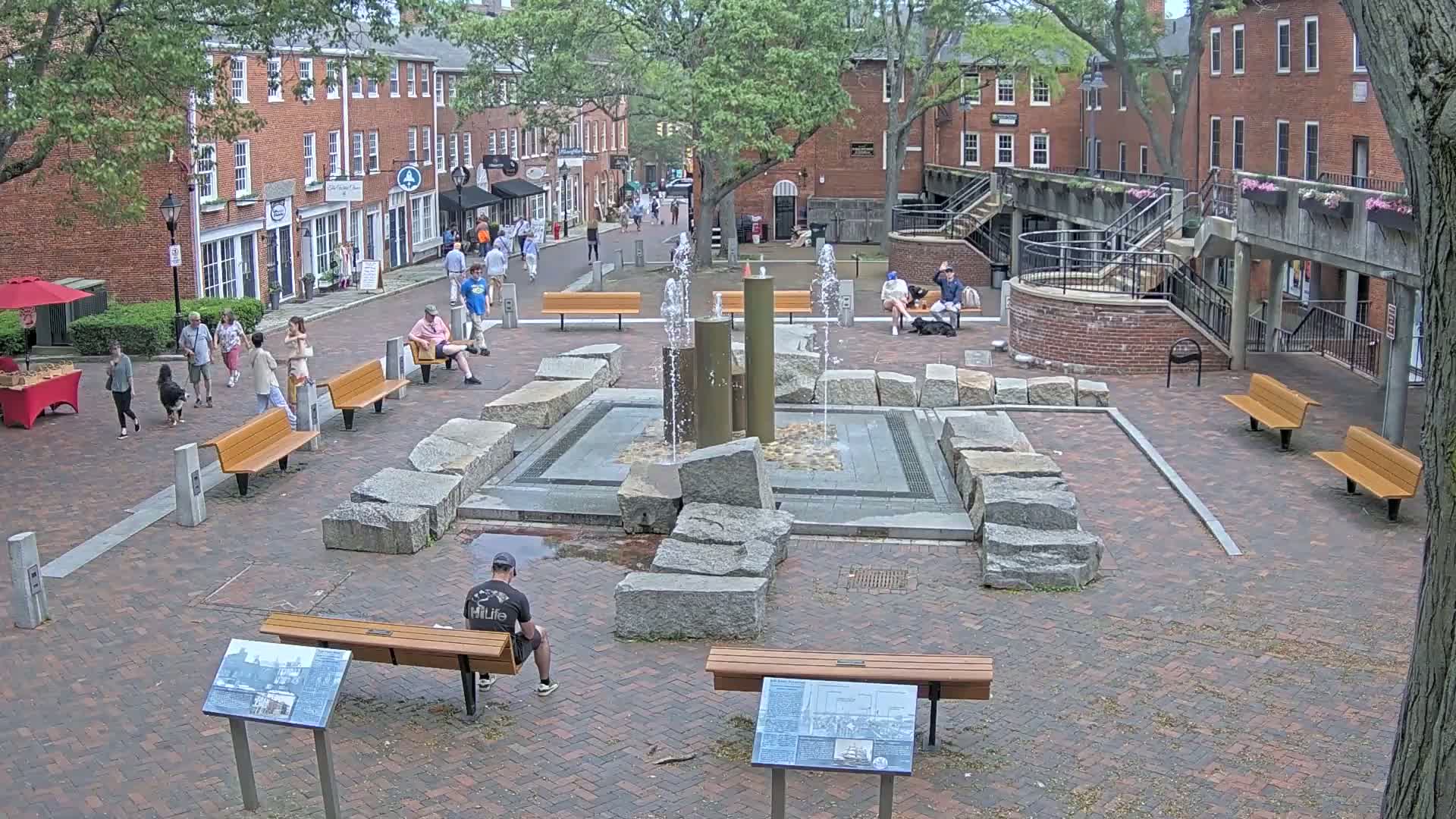 A brick plaza with a central fountain, surrounded by benches, trees, and brick buildings, on a partly sunny day.