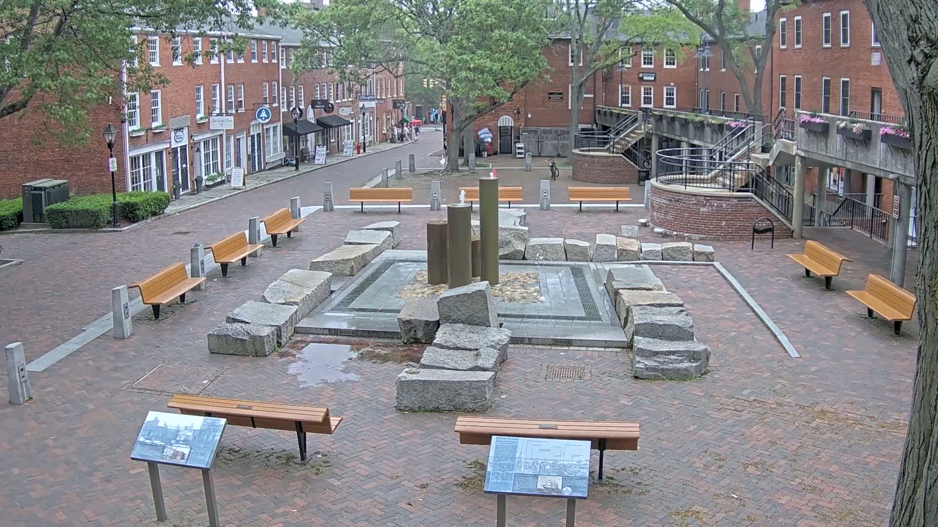 A brick plaza with several benches surrounds a stone fountain, under an overcast sky.