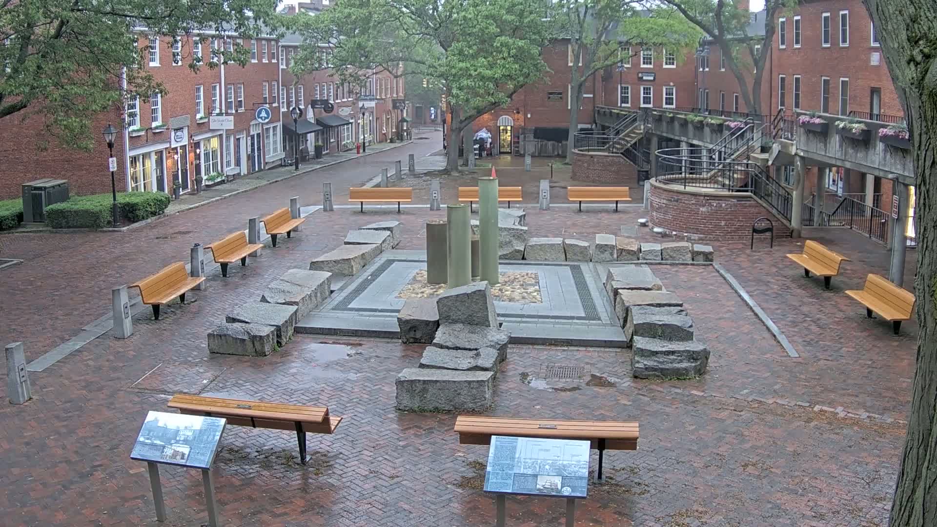 A brick plaza with several benches surrounds a stone and metal fountain, all under an overcast sky.