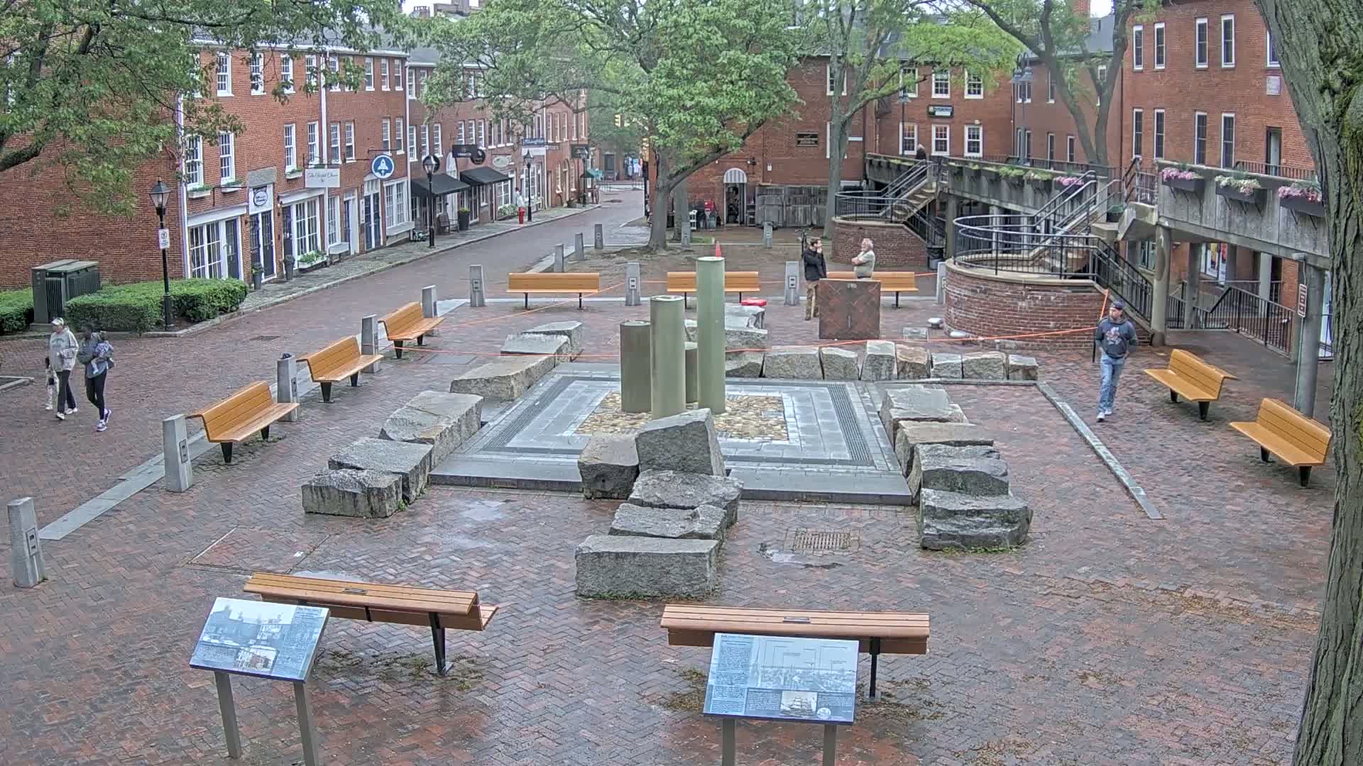 A brick plaza with several benches, a stone fountain, and a few pedestrians under an overcast sky.