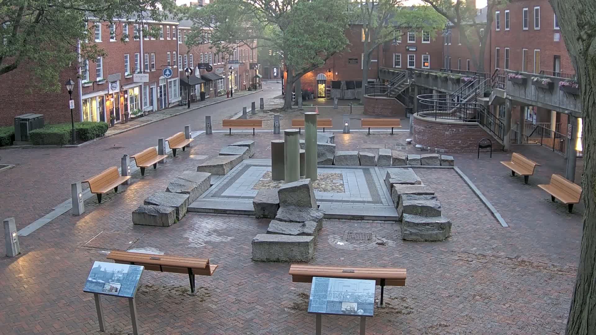 A brick plaza with several benches, a stone fountain, and informational plaques, situated between brick buildings under an overcast sky.