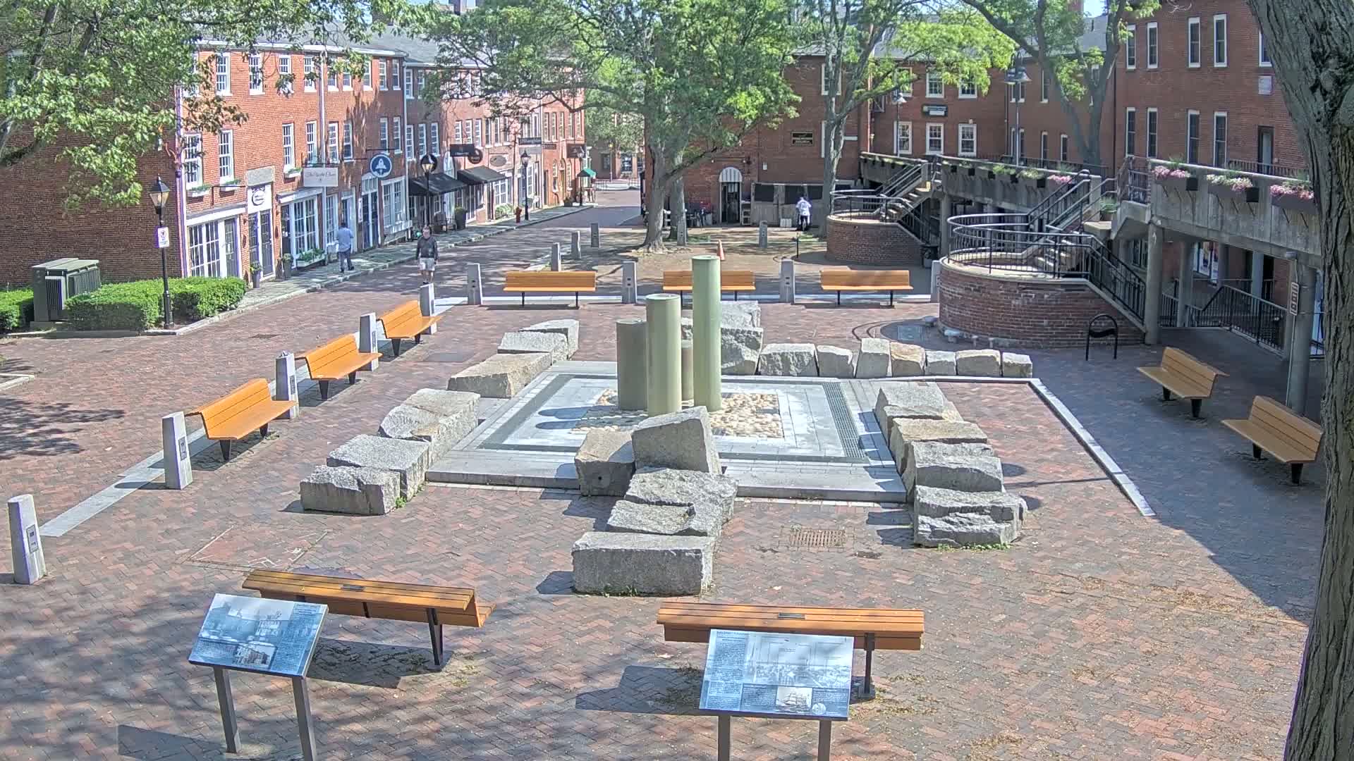 A brick plaza with several benches surrounds a stone fountain, situated between brick buildings under a sunny sky.