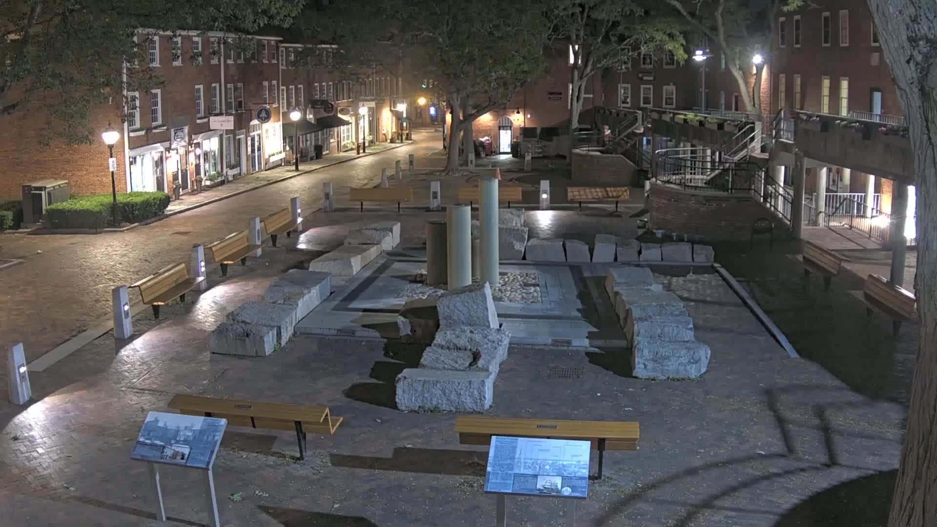 At night, a brick-lined street leads to a plaza featuring large stone ruins and several benches under the shade of trees.