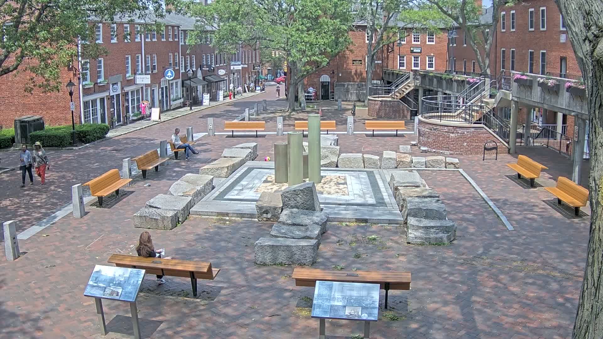 A brick plaza with several benches, a stone fountain, and surrounding brick buildings under a partly sunny sky.