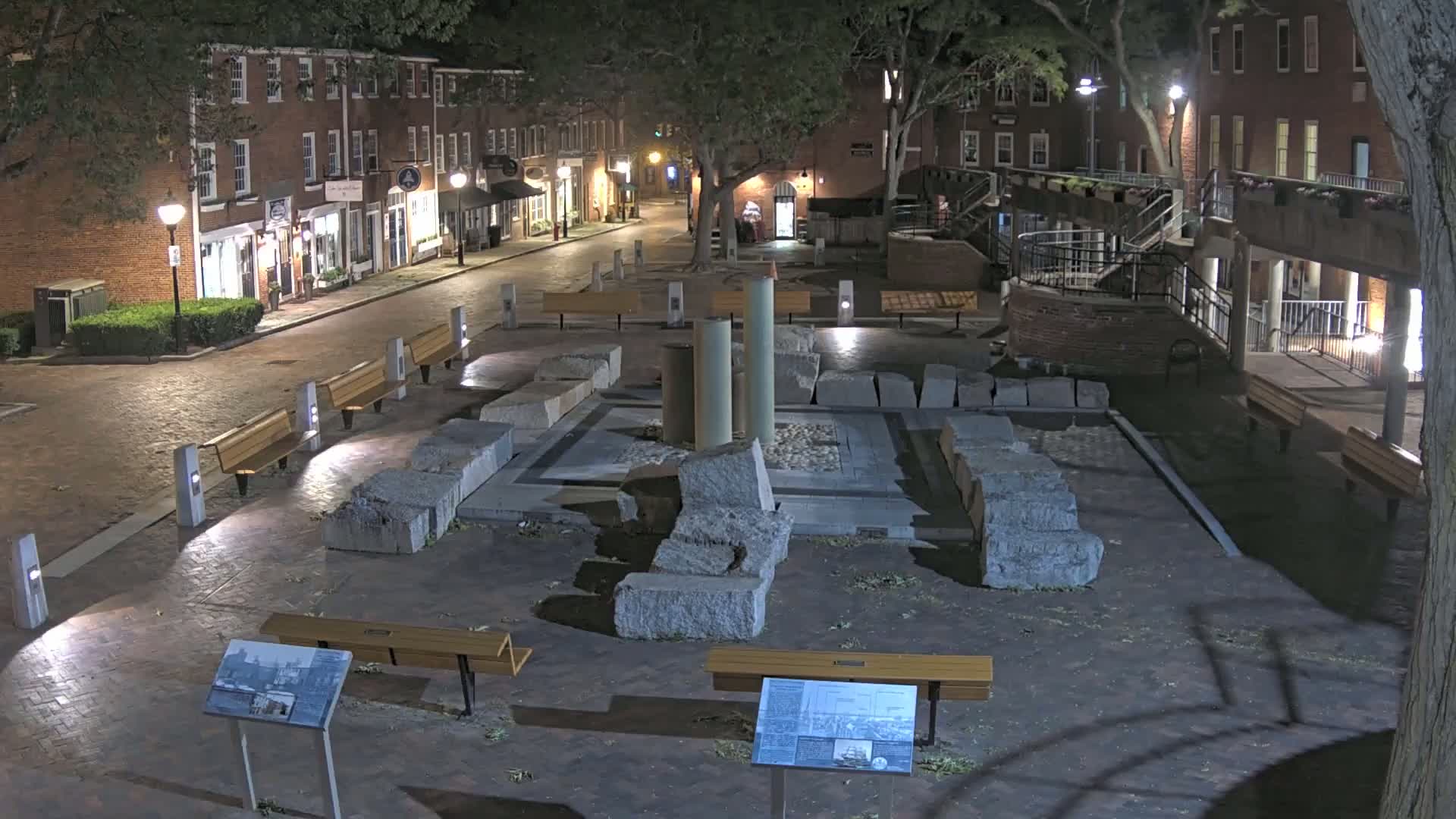 A nighttime view of a town square with a stone monument, benches, and brick buildings lining a street.