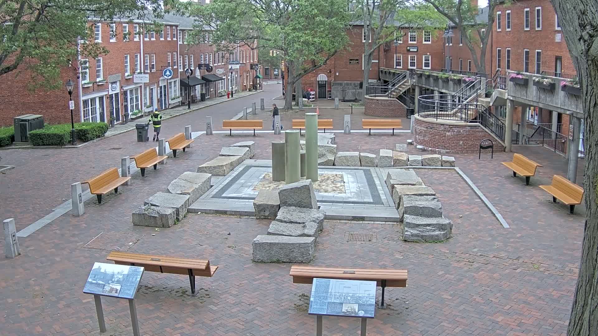 A brick plaza with benches and a stone structure in the center, surrounded by brick buildings under an overcast sky.