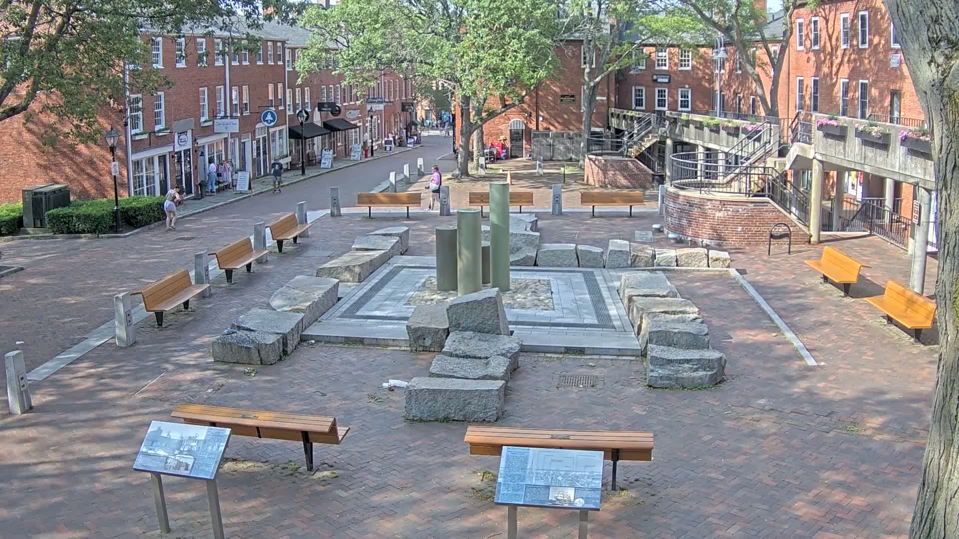 A brick plaza with several benches surrounds a stone fountain-like structure, under a sunny sky.