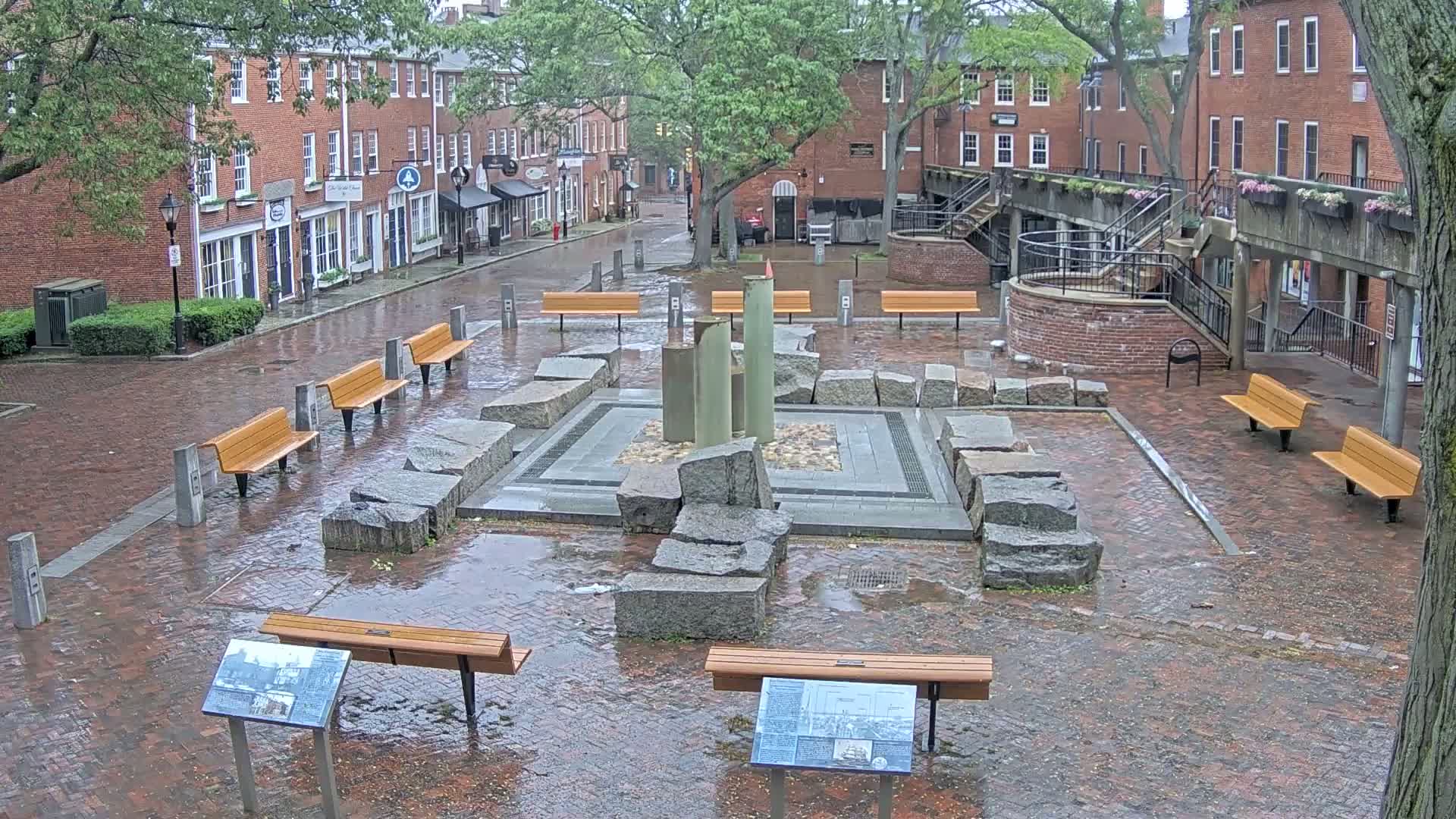 A rain-soaked brick plaza features a central stone fountain and several benches surrounded by red brick buildings.