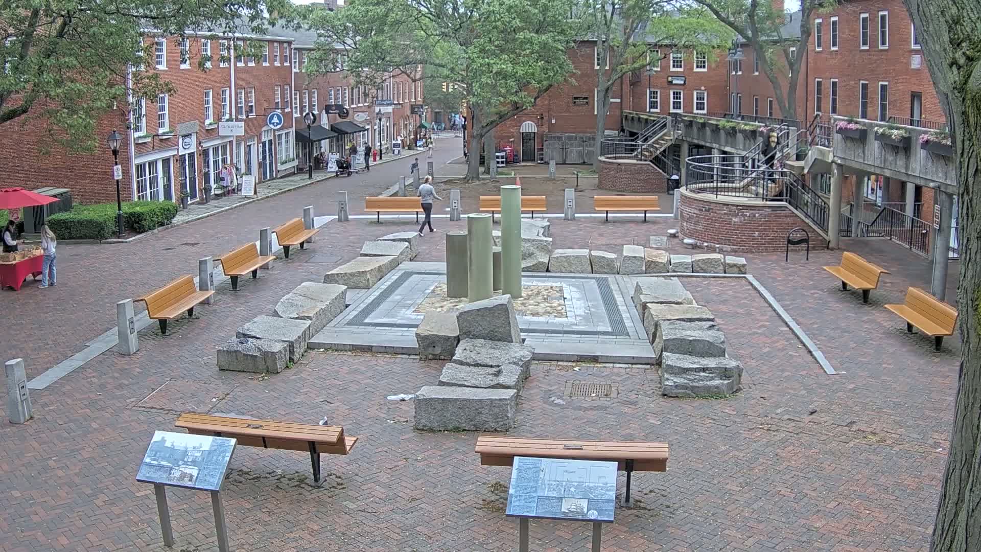 A brick plaza with several benches surrounds a stone fountain-like structure, under an overcast sky.